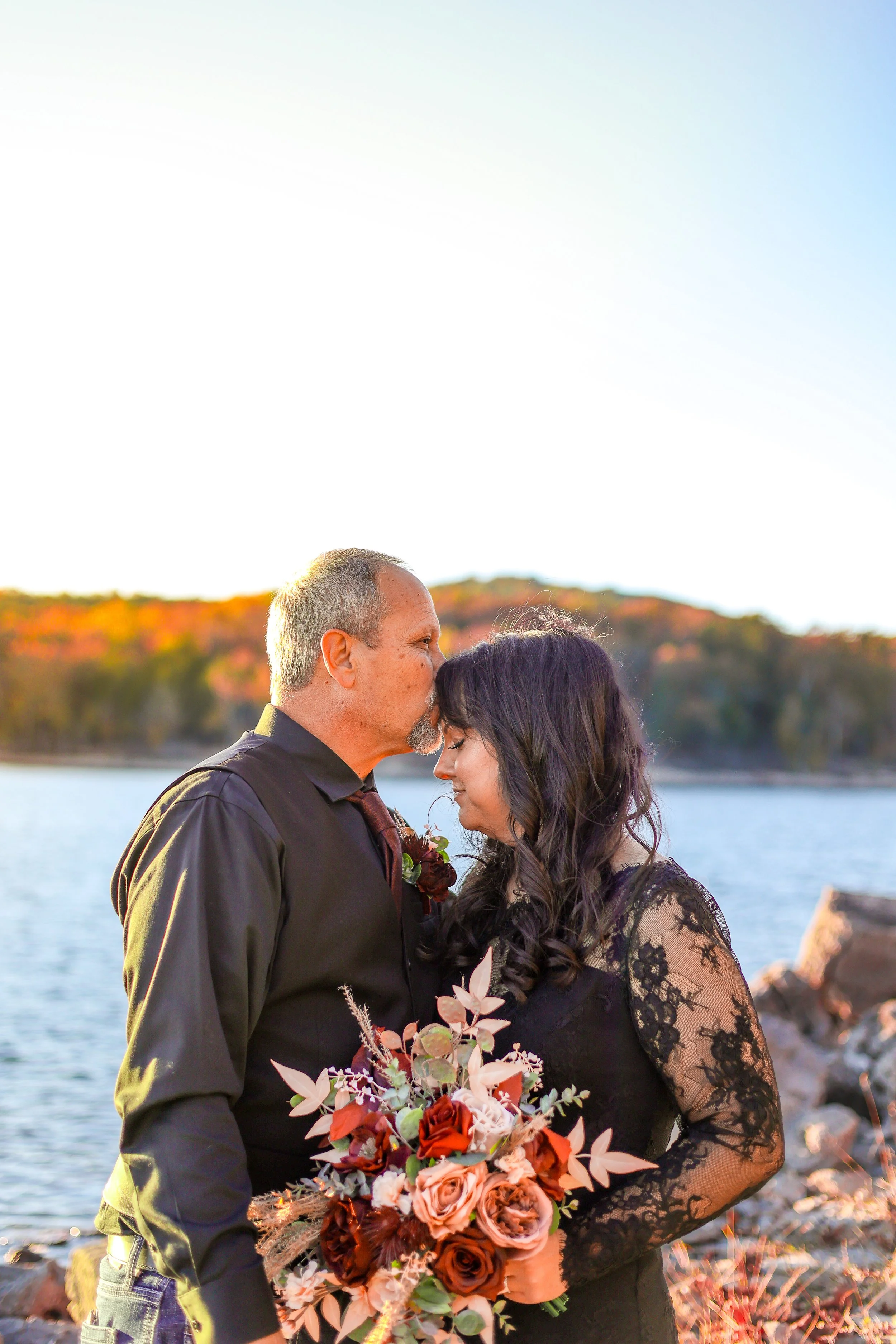 A couple facing each other with foreheads touching, standing outdoors near a body of water with a fall-colored landscape in the background. The man is wearing a black shirt and the woman is dressed in black lace. The woman is holding a large bouquet of flowers with autumnal colors.