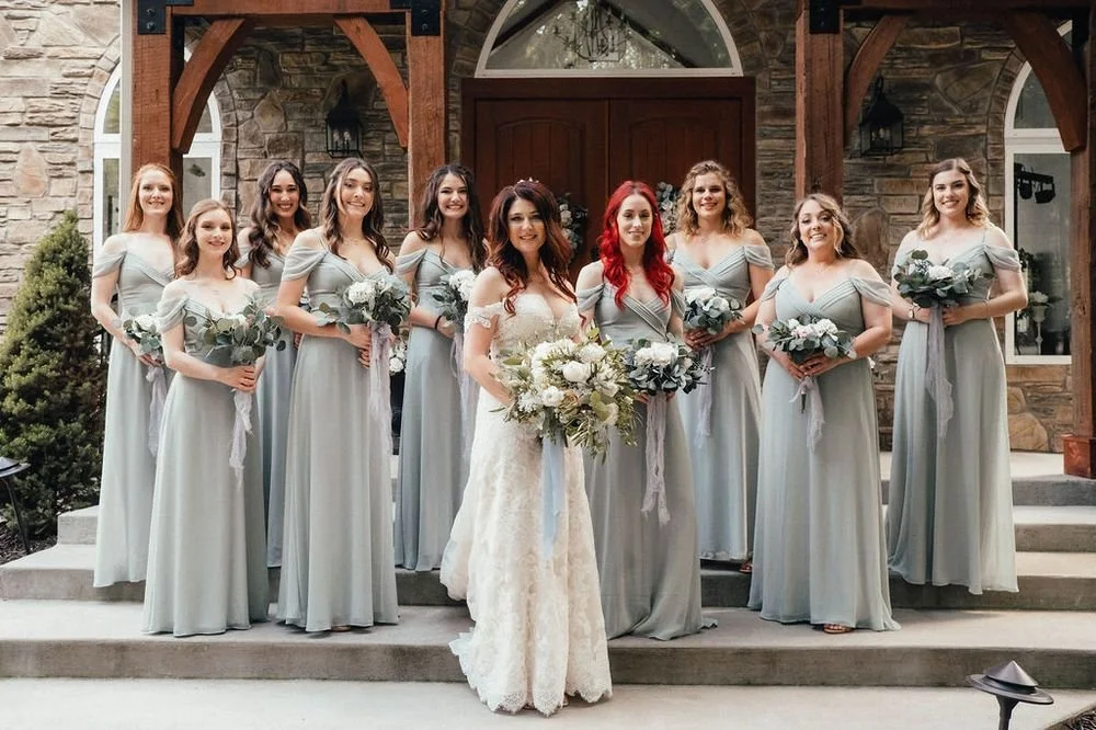 A bride in a white wedding gown standing with her bridesmaids in light blue dresses on the steps in front of a stone and wood building.