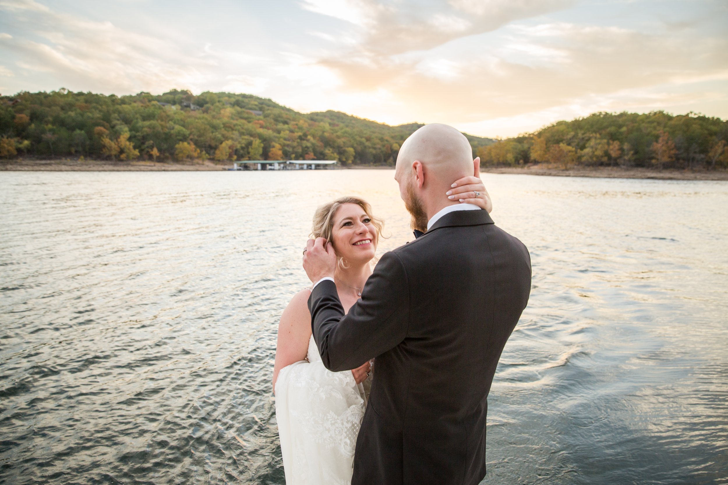 A bride and groom embrace by the water during sunset, with trees on a hillside in the background.