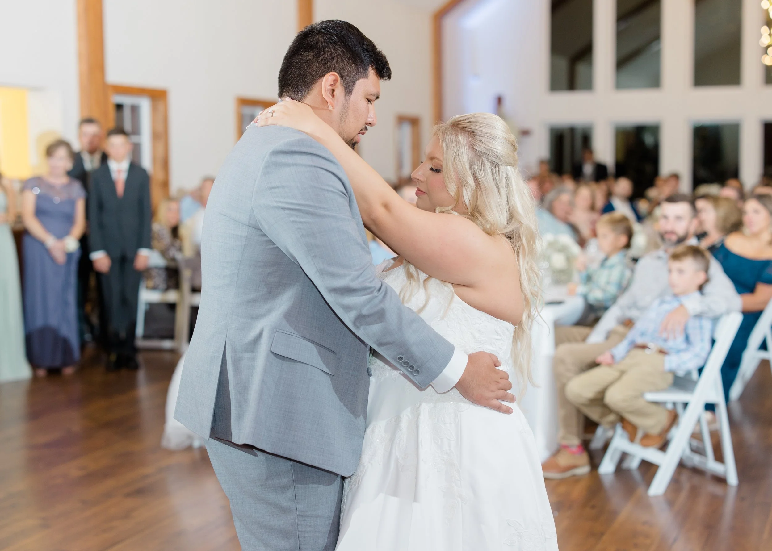A bride and groom share a dance during their wedding reception, surrounded by seated guests in a bright indoor venue.