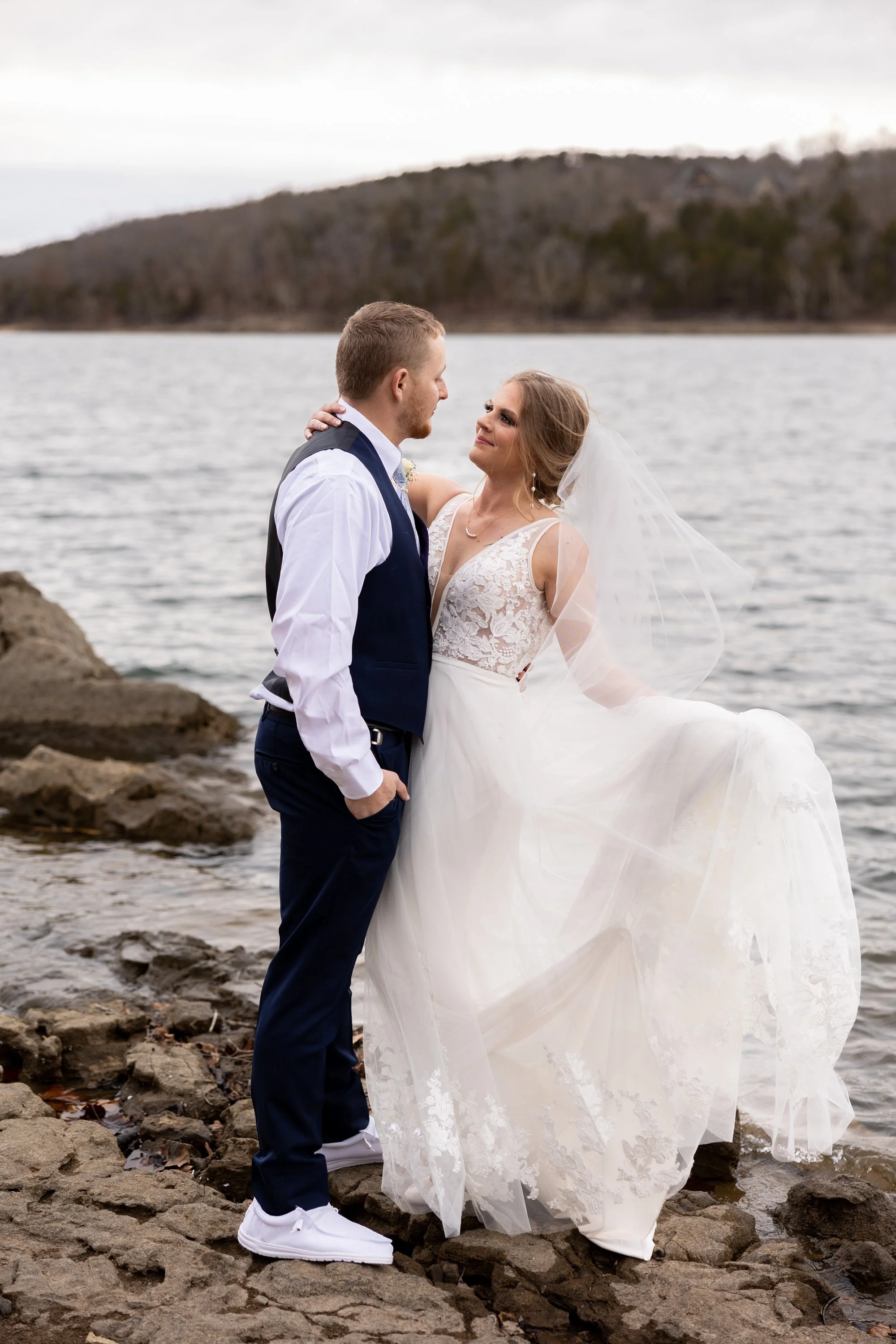 A bride and groom are standing on rocks by a body of water, facing each other, with a tree-covered hill in the background. The groom is wearing a white shirt, navy vest, navy pants, and white sneakers. The bride is wearing a white lace wedding dress with a veil. They are holding each other and smiling at each other.