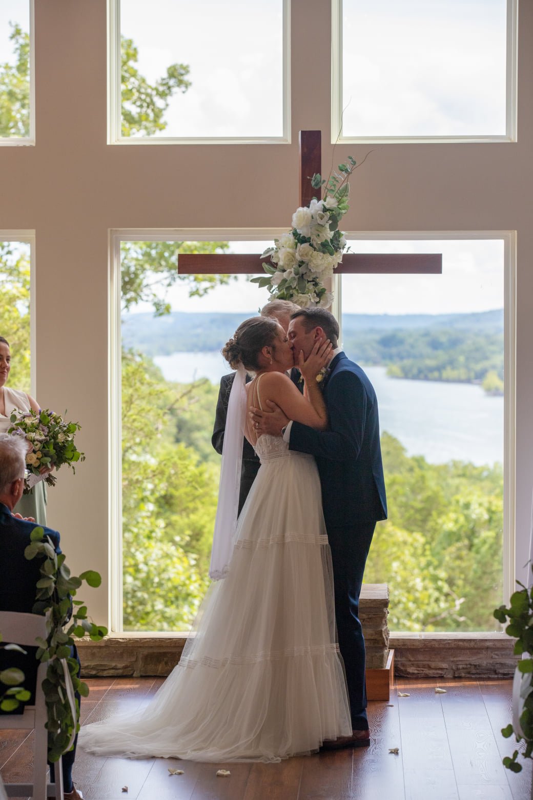 A wedding ceremony with a bride and groom kissing in front of a large window with a scenic lake view, decorated with a cross and white flowers.