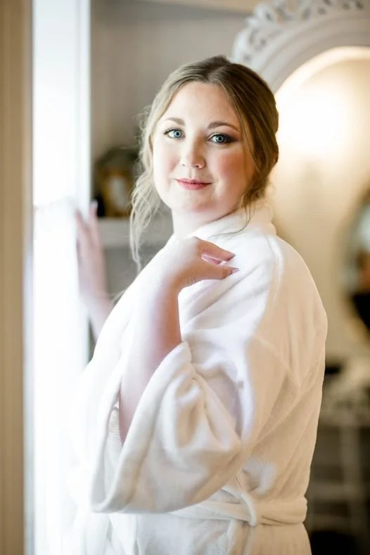 A woman with light skin, blonde hair, and blue eyes standing indoors near a window, wearing a white bathrobe and smiling at the camera.