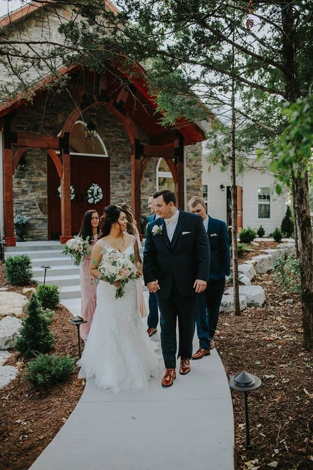 A wedding party walking on a garden pathway outside a rustic church during daytime. The bride and groom are holding hands, with the bride holding a bouquet of white flowers. The bride wears a white wedding gown, and the groom wears a dark blue suit. 