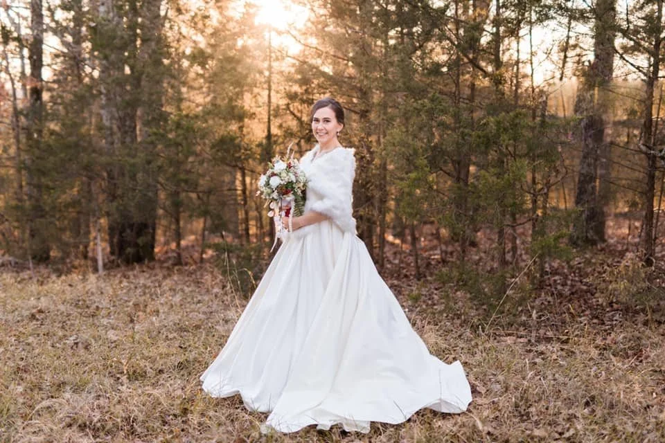 A bride in a white wedding dress holding a bouquet of flowers, standing outdoors in a wooded area during sunset.