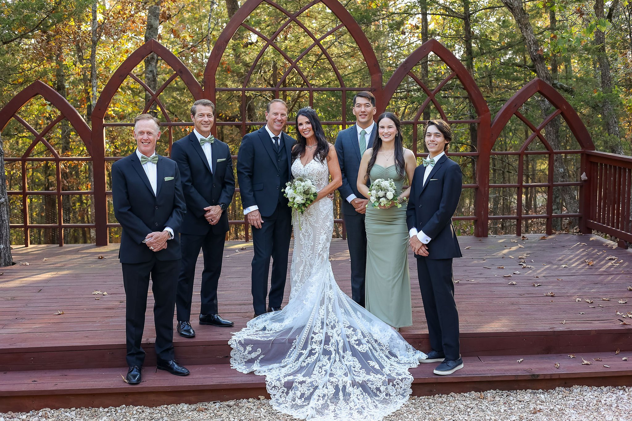 A wedding party of seven people standing on a wooden deck outdoors with trees in the background. The bride is in the center wearing a white lace wedding gown holding a bouquet, surrounded by five men in suits with bow ties and a woman in a sage green