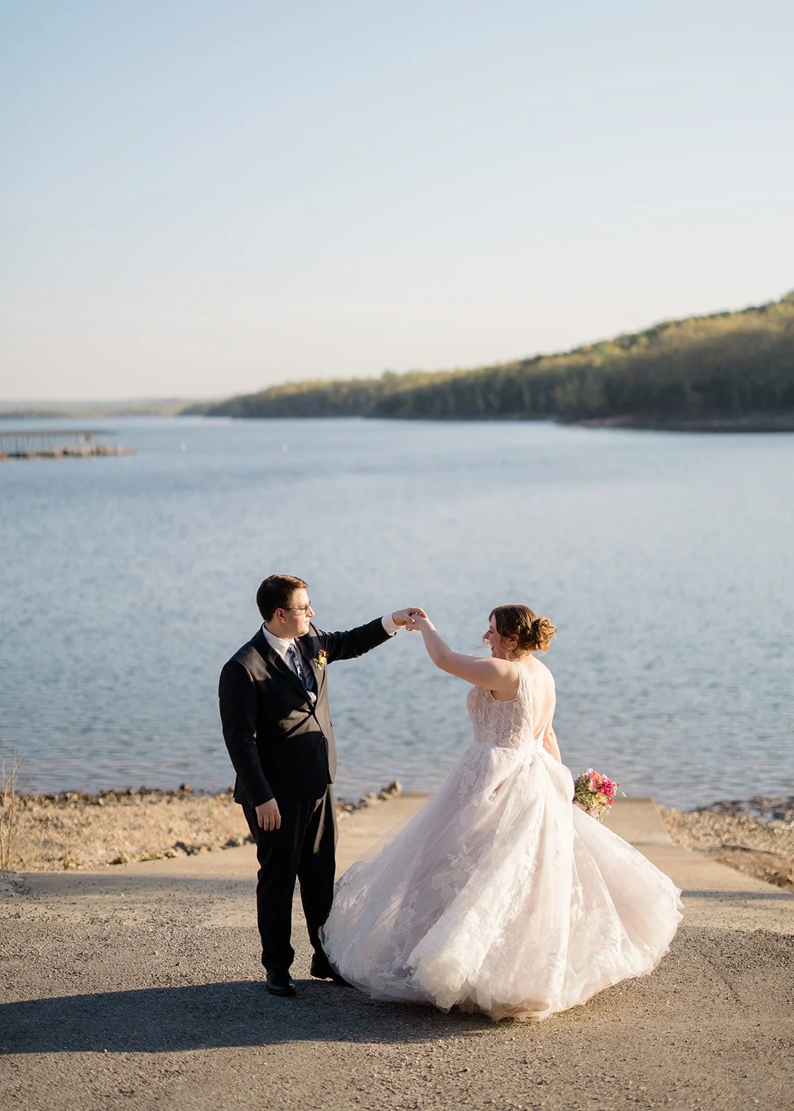 A bride and groom dancing by a lake with hills in the background, on their wedding day; the bride wears a white wedding gown and the groom a black suit.