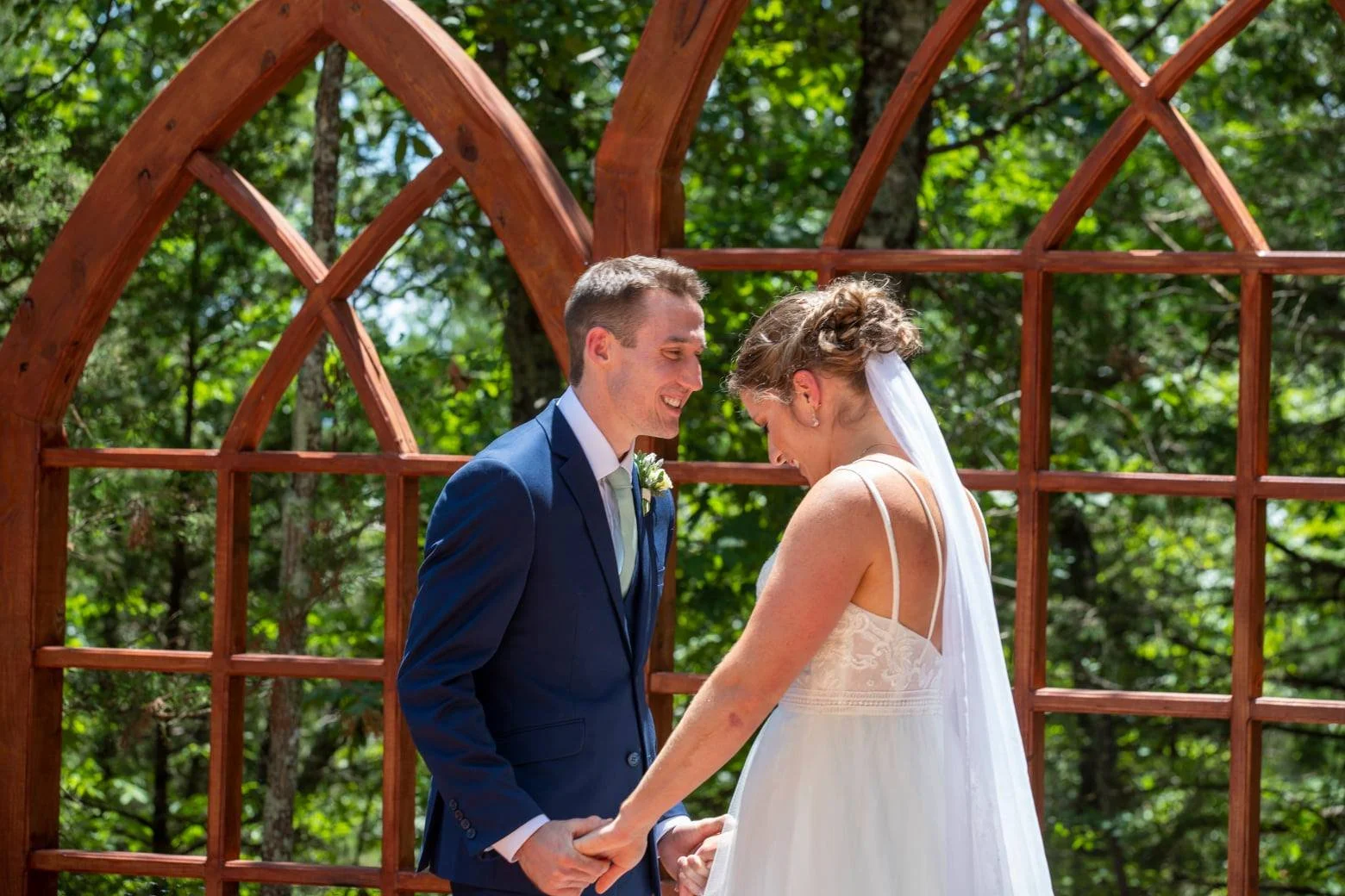A bride and groom holding hands and smiling during their outdoor wedding ceremony, with wooden arches and lush green trees in the background.