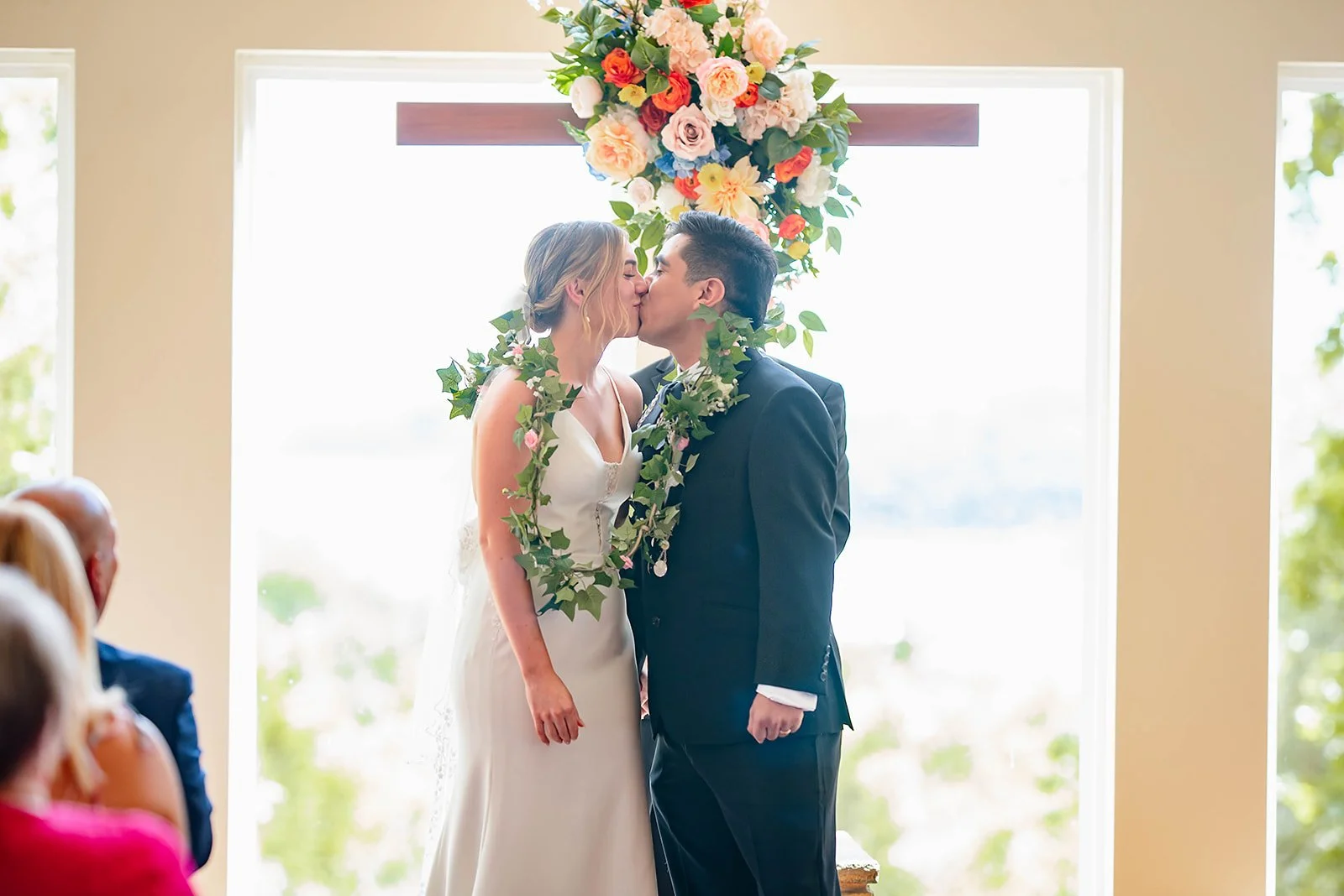 A bride and groom kiss during their wedding ceremony under a floral arch, with sunlight shining through large windows behind them.