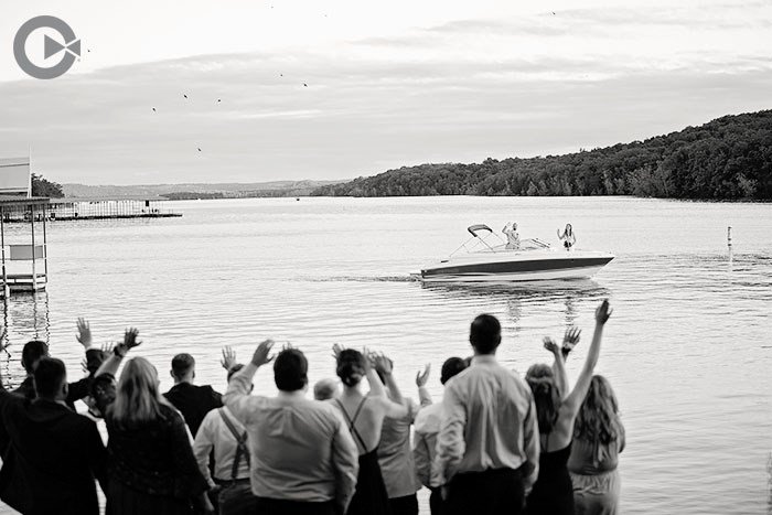 Group of people waving towards a boat on a lake, with a shoreline and trees in the background