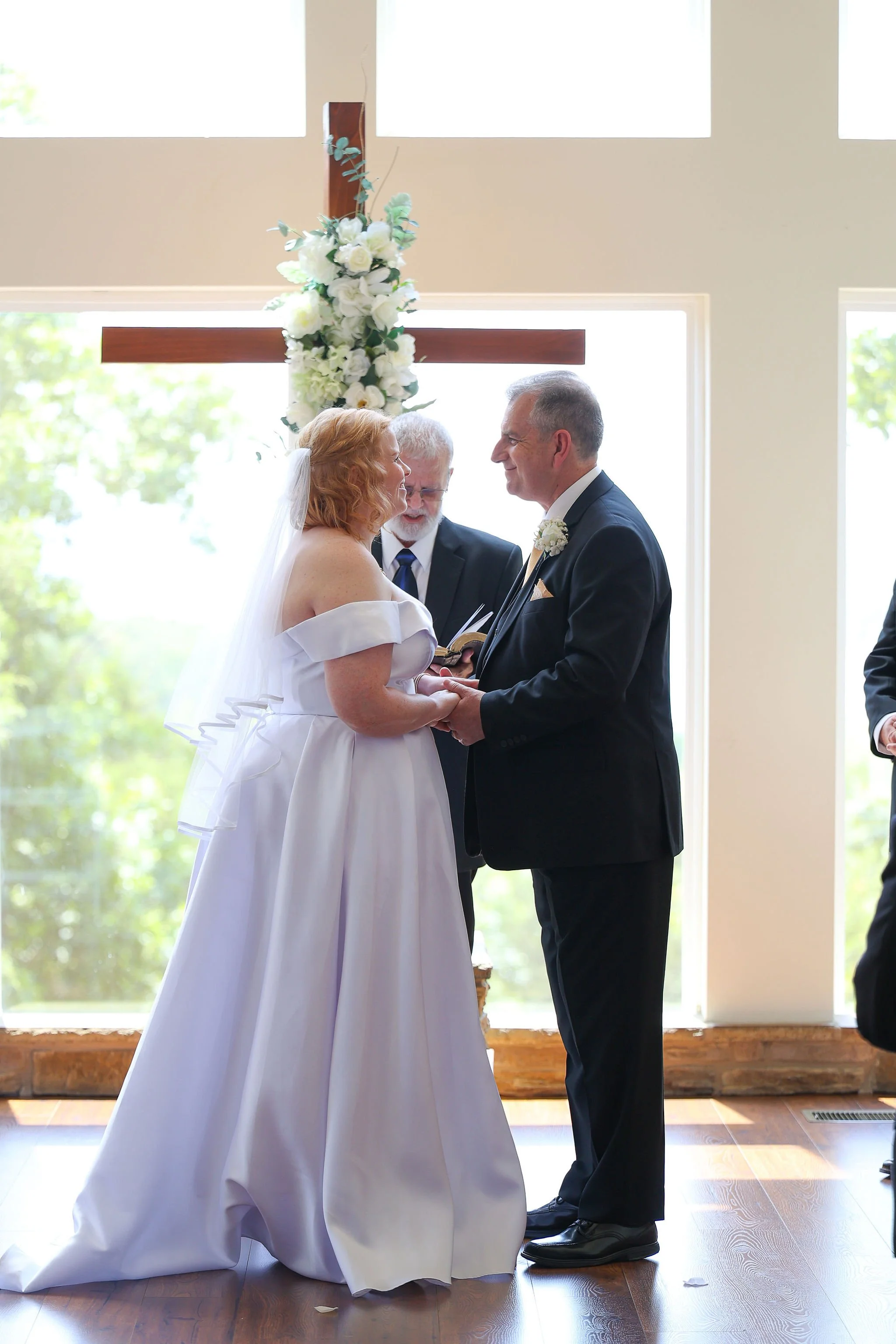 A bride and groom holding hands and smiling at each other during their wedding ceremony inside a church with a large cross decorated with flowers in the background.