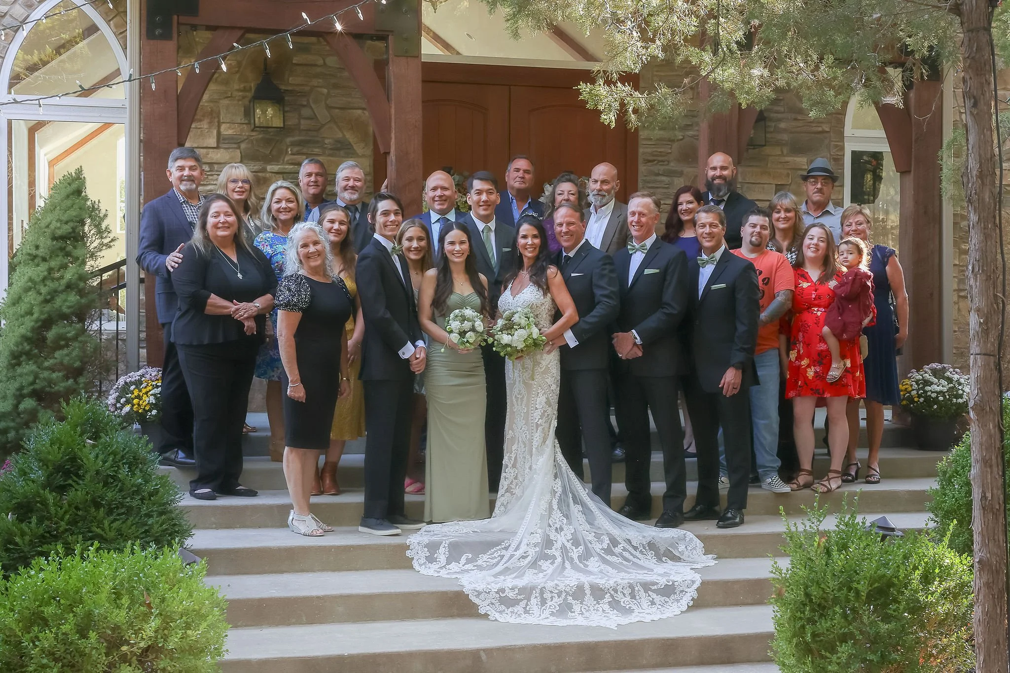 A large group of people, including men, women, children, and a bride and groom, standing on outdoor steps in front of a stone building with trees and greenery around. The bride is wearing a white lace wedding dress, holding a bouquet, and the groom is in a tuxedo with a bow tie.