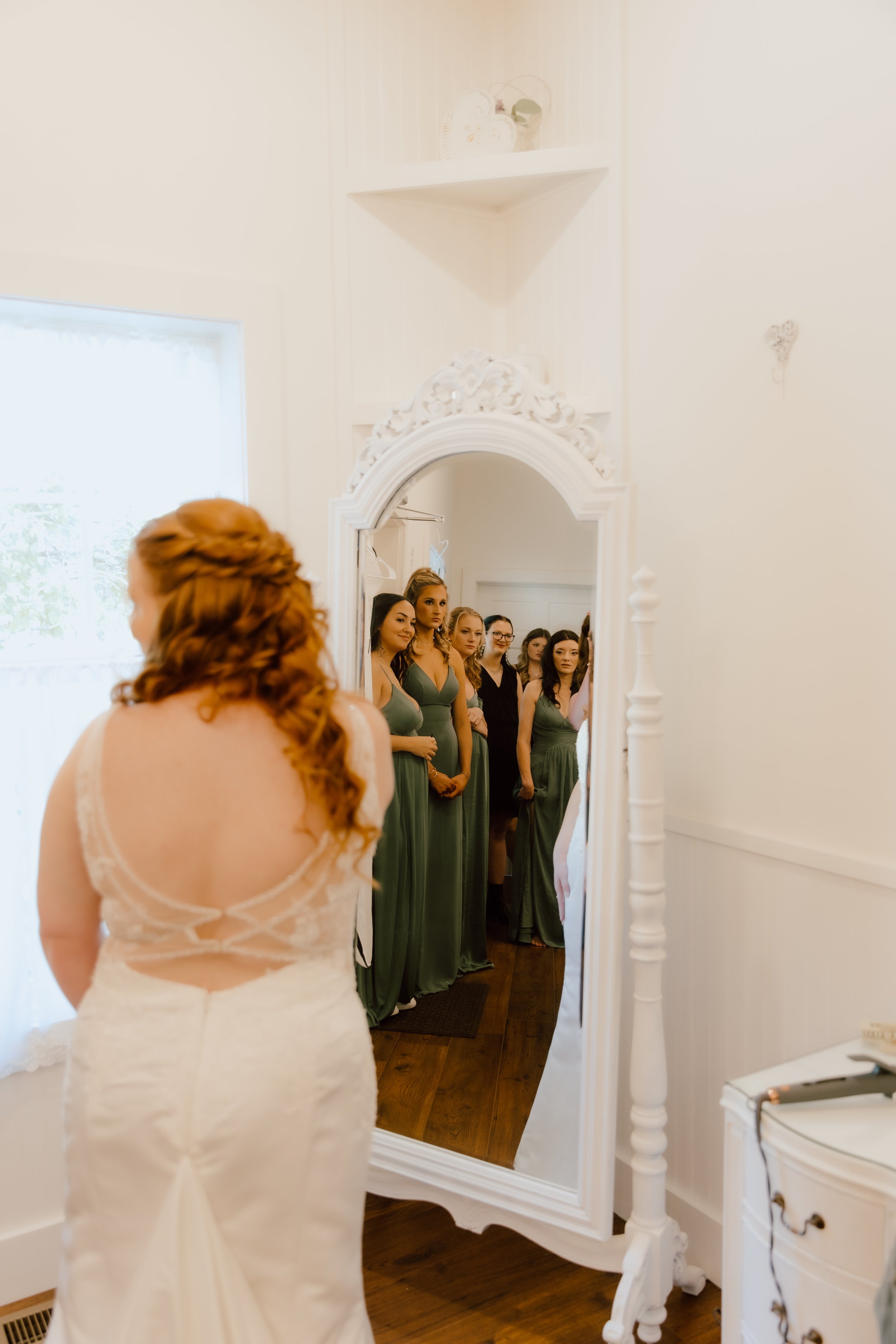 A bride with red hair in a white lace wedding gown facing a mirror, surrounded by her bridesmaids in green dresses.