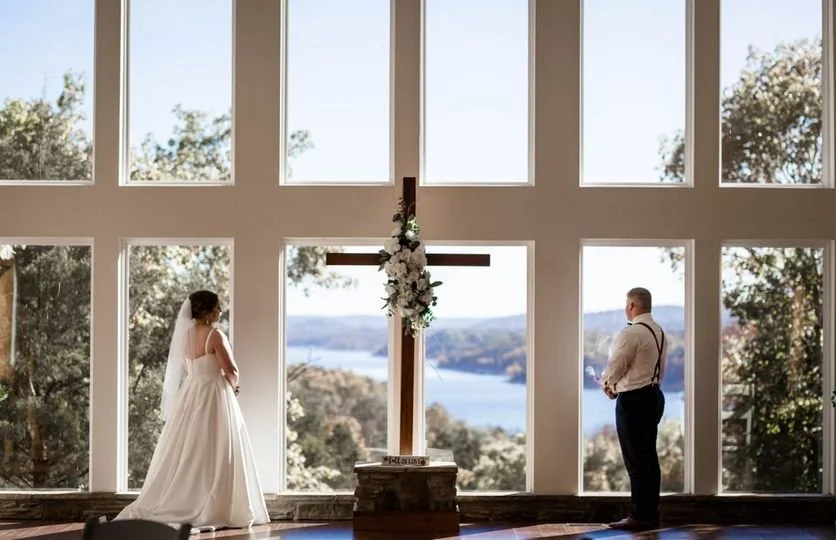 Bride and groom standing apart in front of a large window with a cross decorated with flowers between them, overlooking a scenic lake and trees.