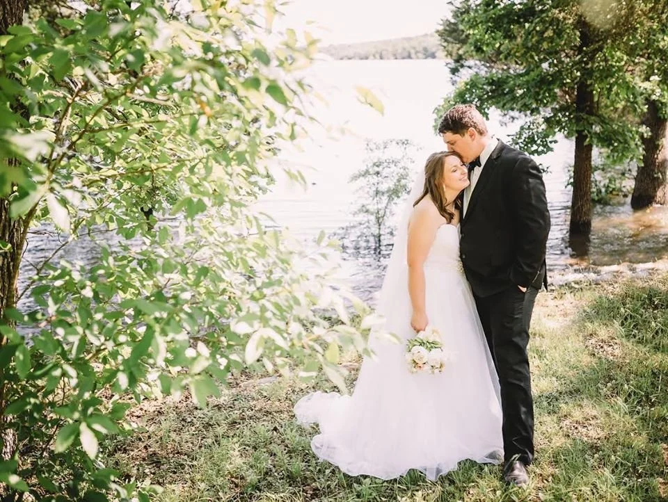 A newlywed couple standing near a lake, embracing each other, with trees in the background on a sunny day.