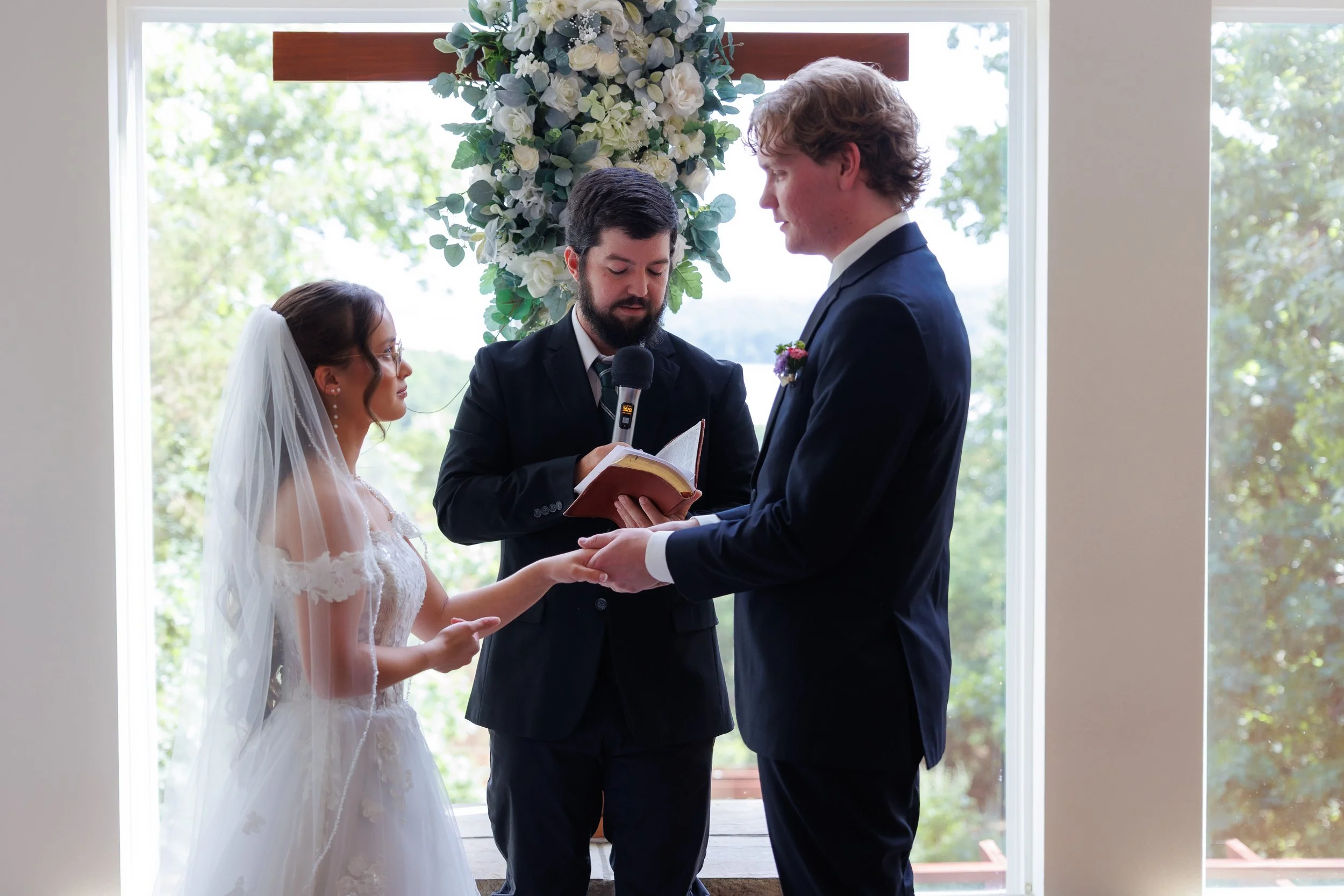A bride and groom holding hands during their wedding ceremony in front of a window with a floral arrangement above them, officiant standing between them reading wedding vows.