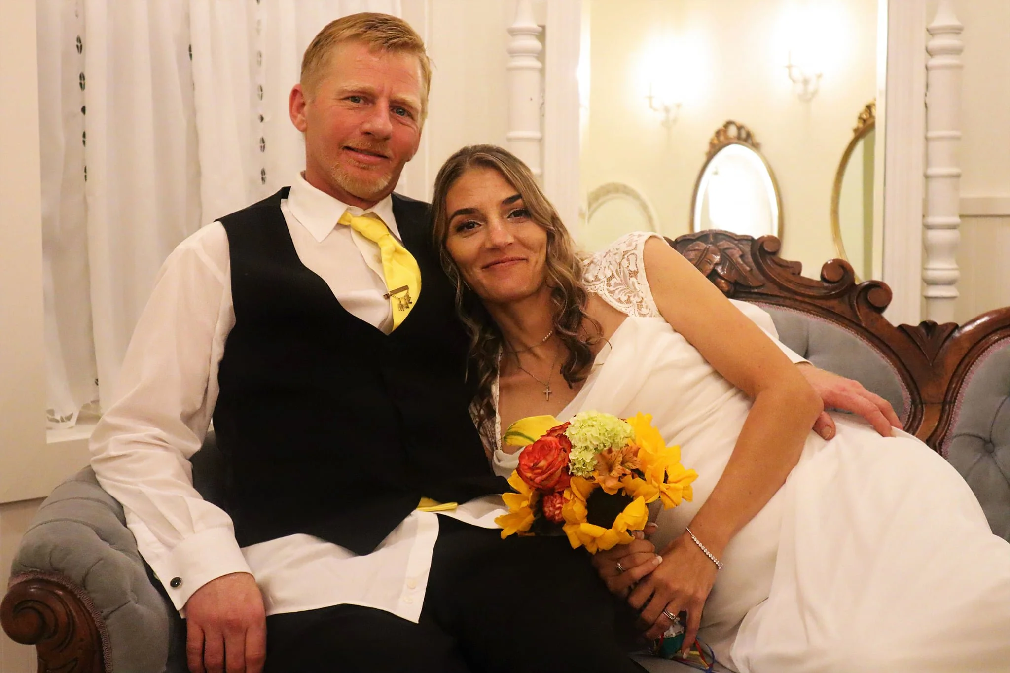 A man and woman sitting on a vintage sofa, the woman holding a bouquet of colorful flowers. The man has short blonde hair and a beard, wearing a white shirt, black vest, and yellow tie. The woman has curly brown hair, wearing a white dress with lace details and a cross necklace. The background shows a well-lit, elegant room with light-colored curtains and decorative mirrors.