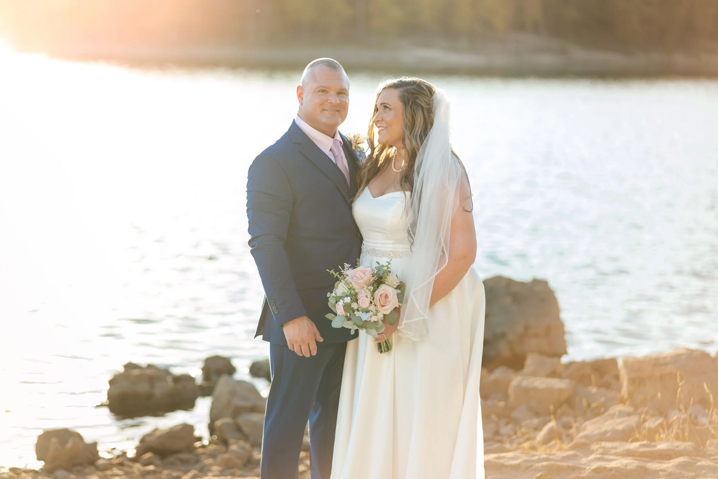 A couple dressed in wedding attire standing on a rocky beach near water, with the sunset in the background, celebrating their wedding.