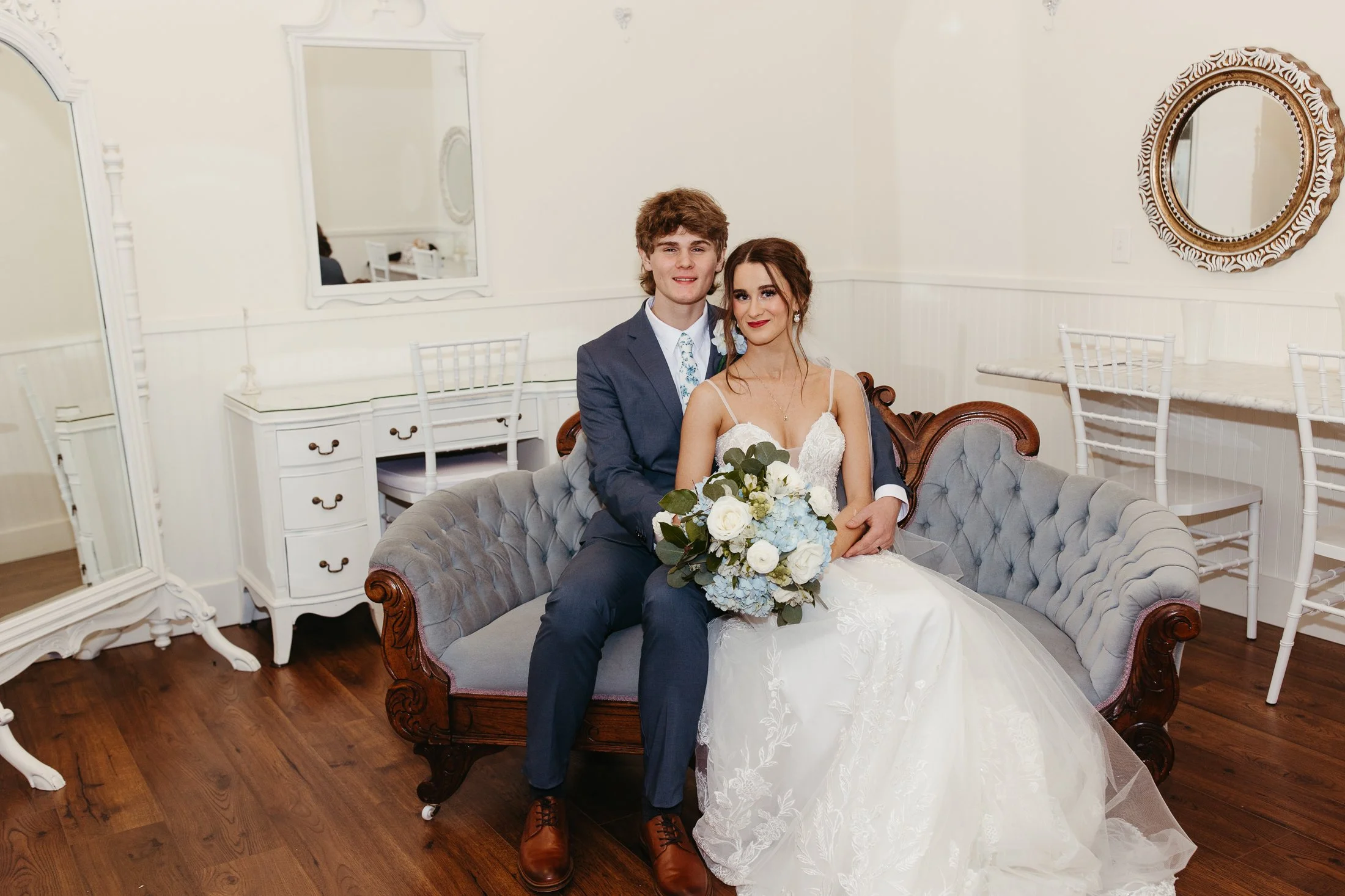 A young couple, dressed in wedding attire, sitting on a vintage-style sofa in a bright, elegant room holding a bouquet of white and blue flowers, smiling at the camera.