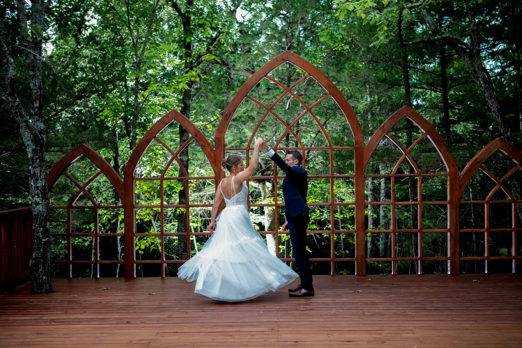 Bride and groom dance together outside on wooden platform with a decorative wooden arch and lush green trees in the background.