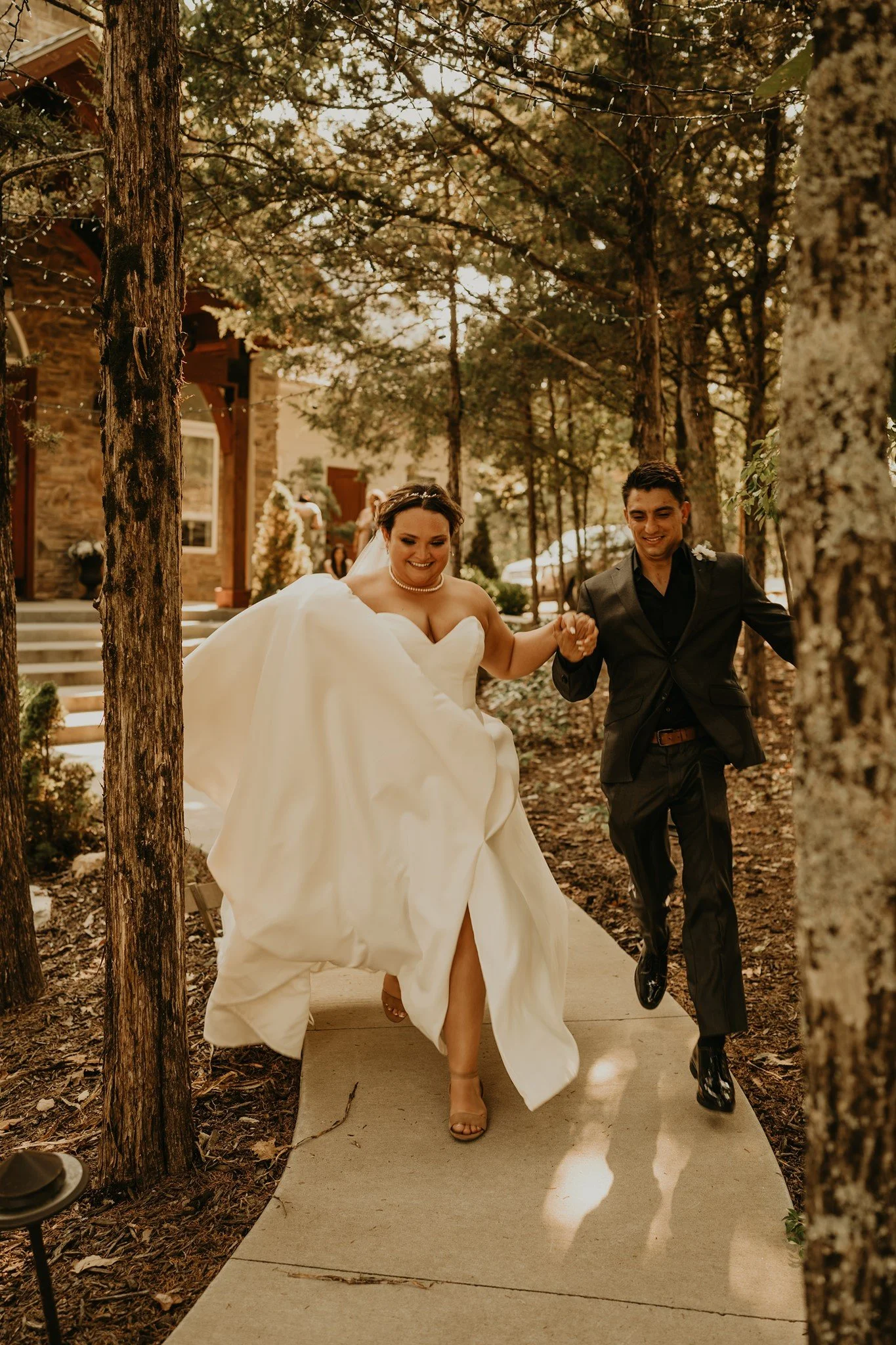 A bride and groom are holding hands and walking outdoors on a sunny day, surrounded by trees, with the bride in a white wedding gown and the groom in a black suit.