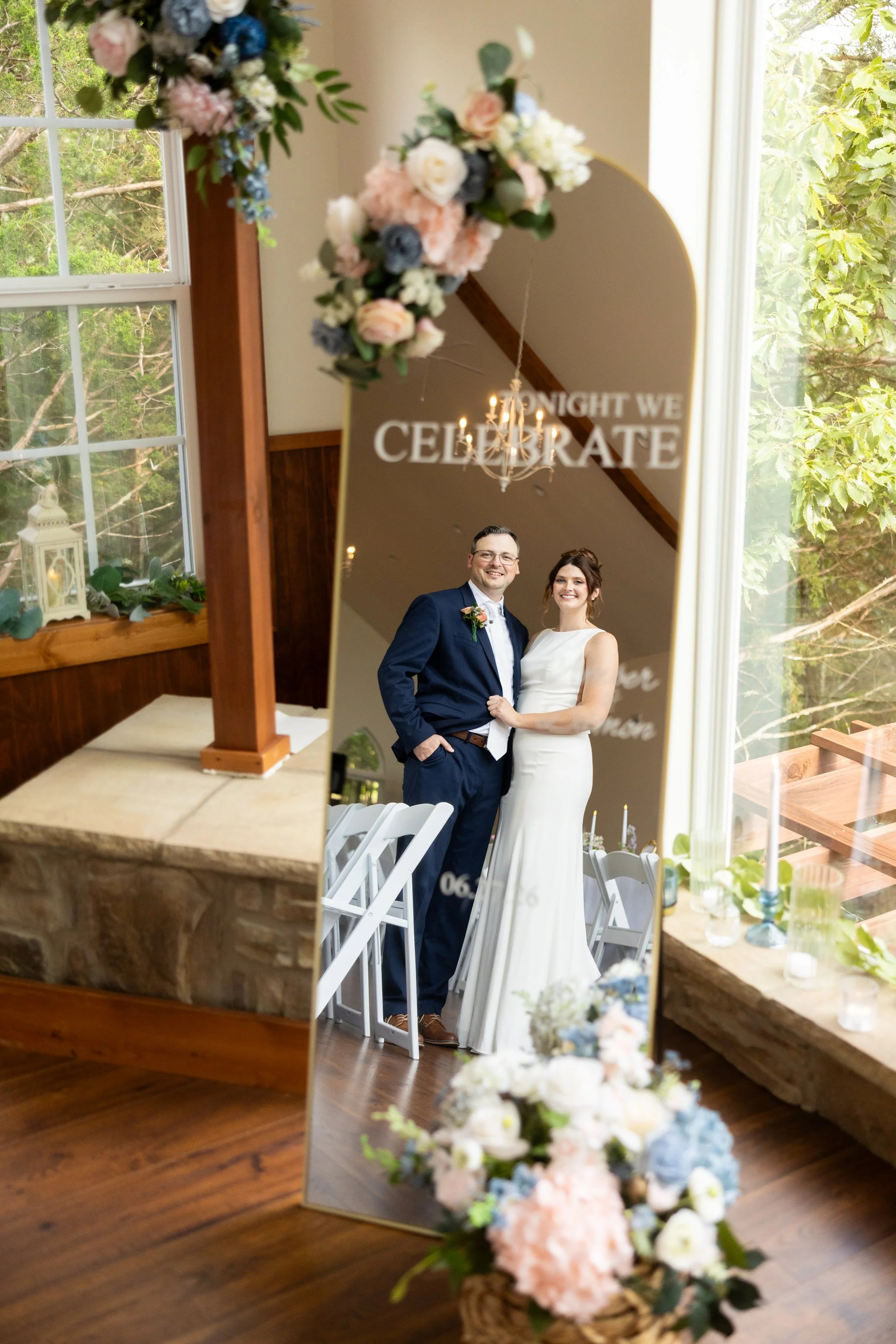A bride and groom smiling in wedding attire are seen through a decorative mirror at a wedding reception, with floral arrangements and wedding decorations around.