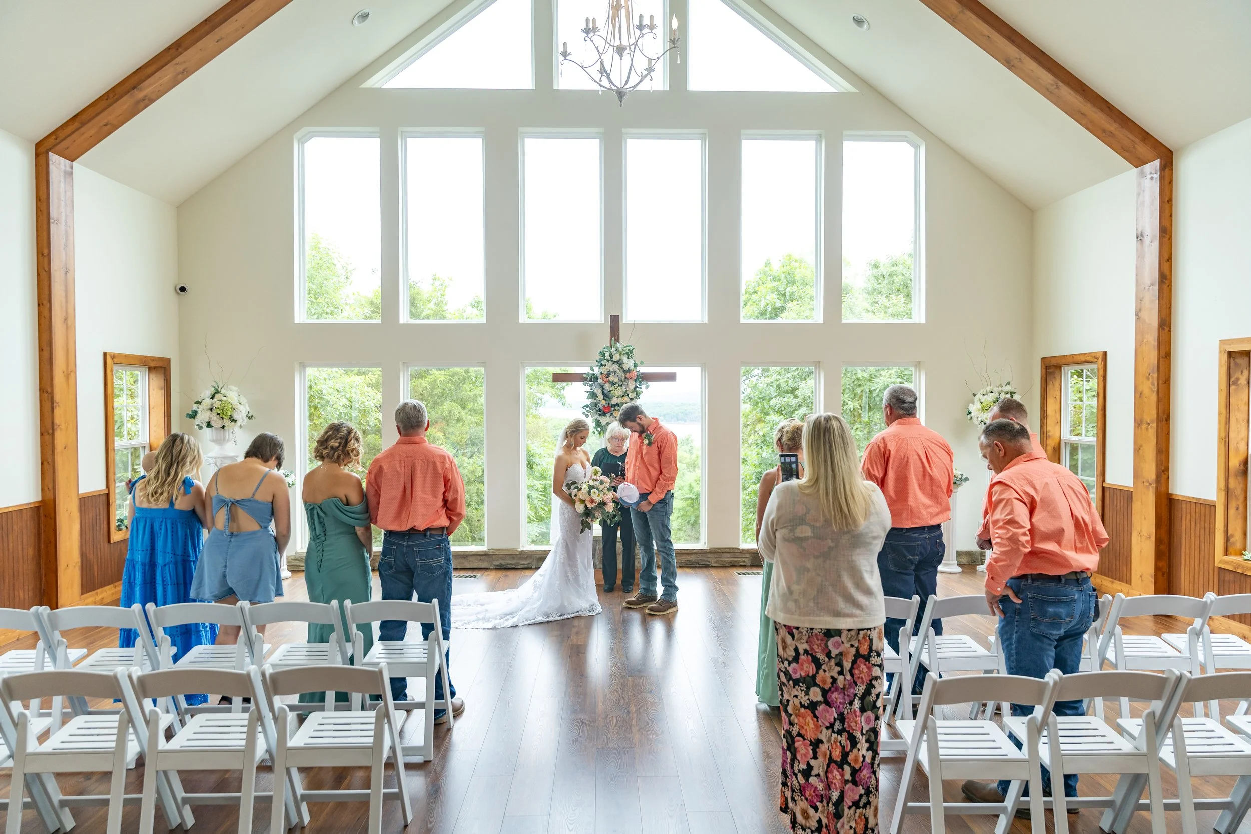 Indoor wedding ceremony with a couple exchanging vows at the altar, surrounded by guests, in a bright room with large windows, wooden beams, and floral decorations.