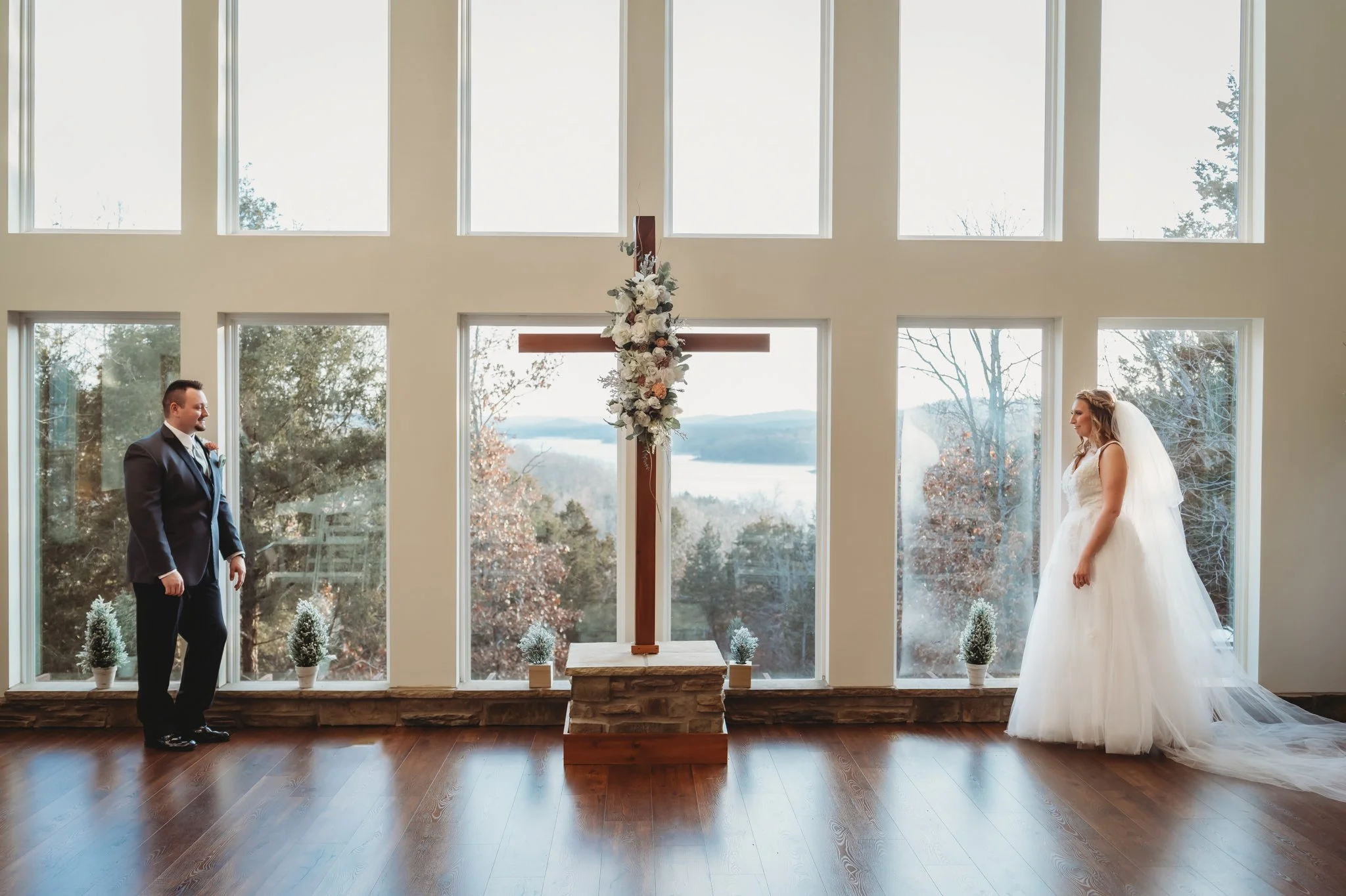 A bride and groom stand apart inside a church with large windows, a cross decorated with flowers at the altar, and scenic mountains outside