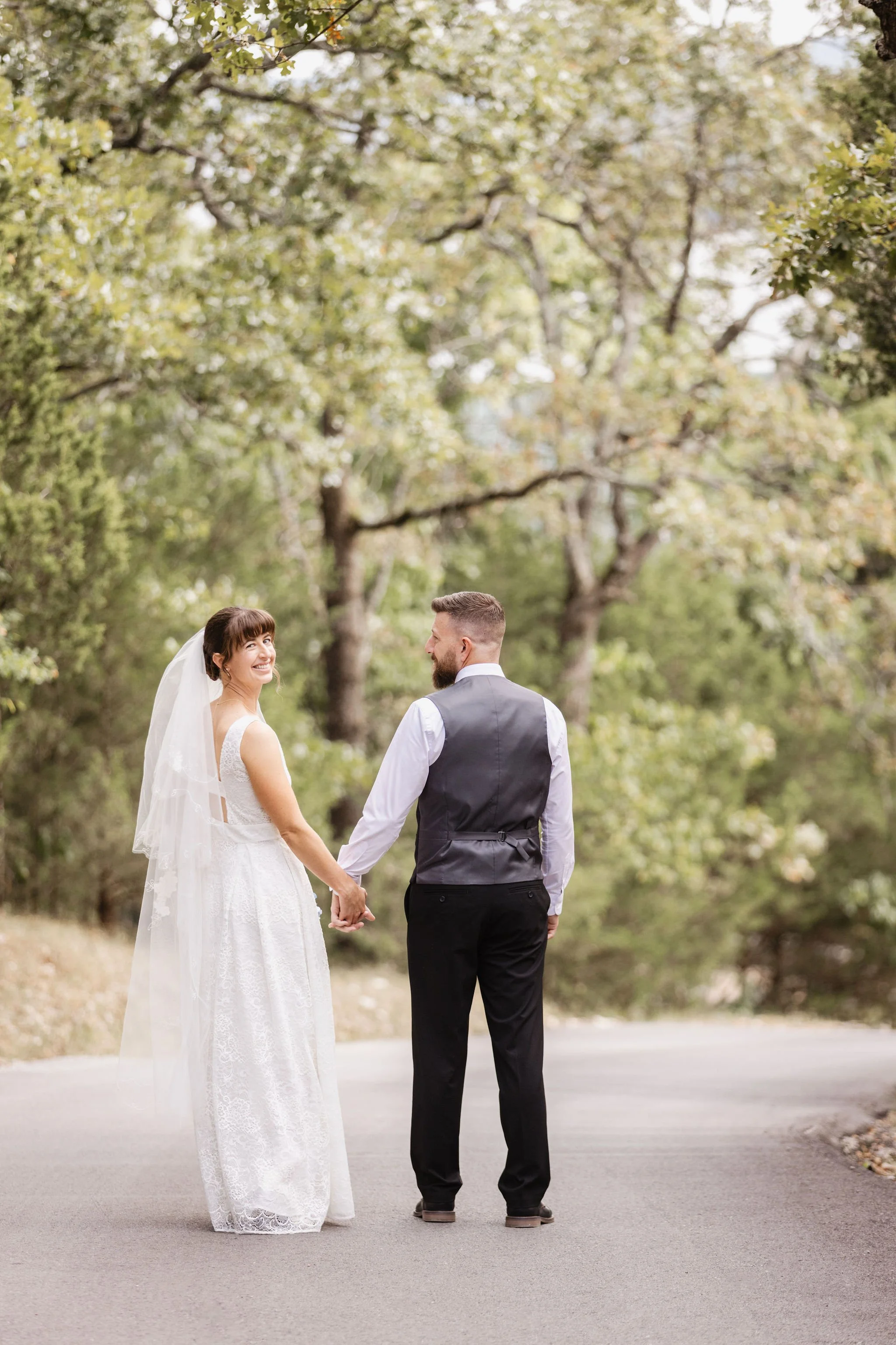 A bride and groom holding hands and smiling at each other outdoors, surrounded by trees with green leaves, on a paved path.