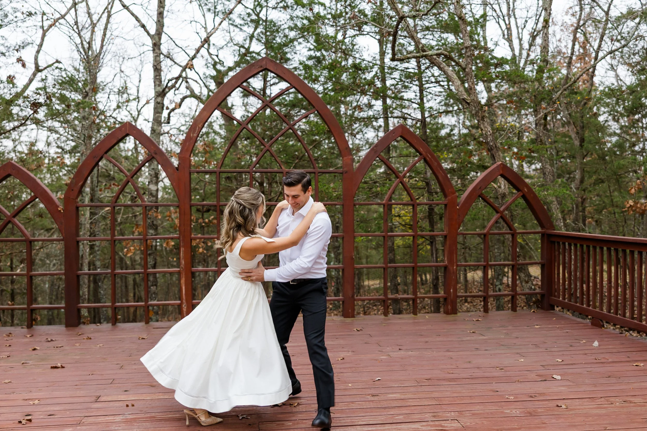 A couple in wedding attire dancing on a wooden deck outdoors, surrounded by trees, with a decorative metal arch in the background.