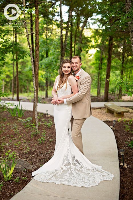 A newlywed couple standing on a curved pathway in a lush green wooded area, smiling at the camera during their wedding photo shoot. The bride is wearing a white lace wedding gown, and the groom is dressed in a beige suit.
