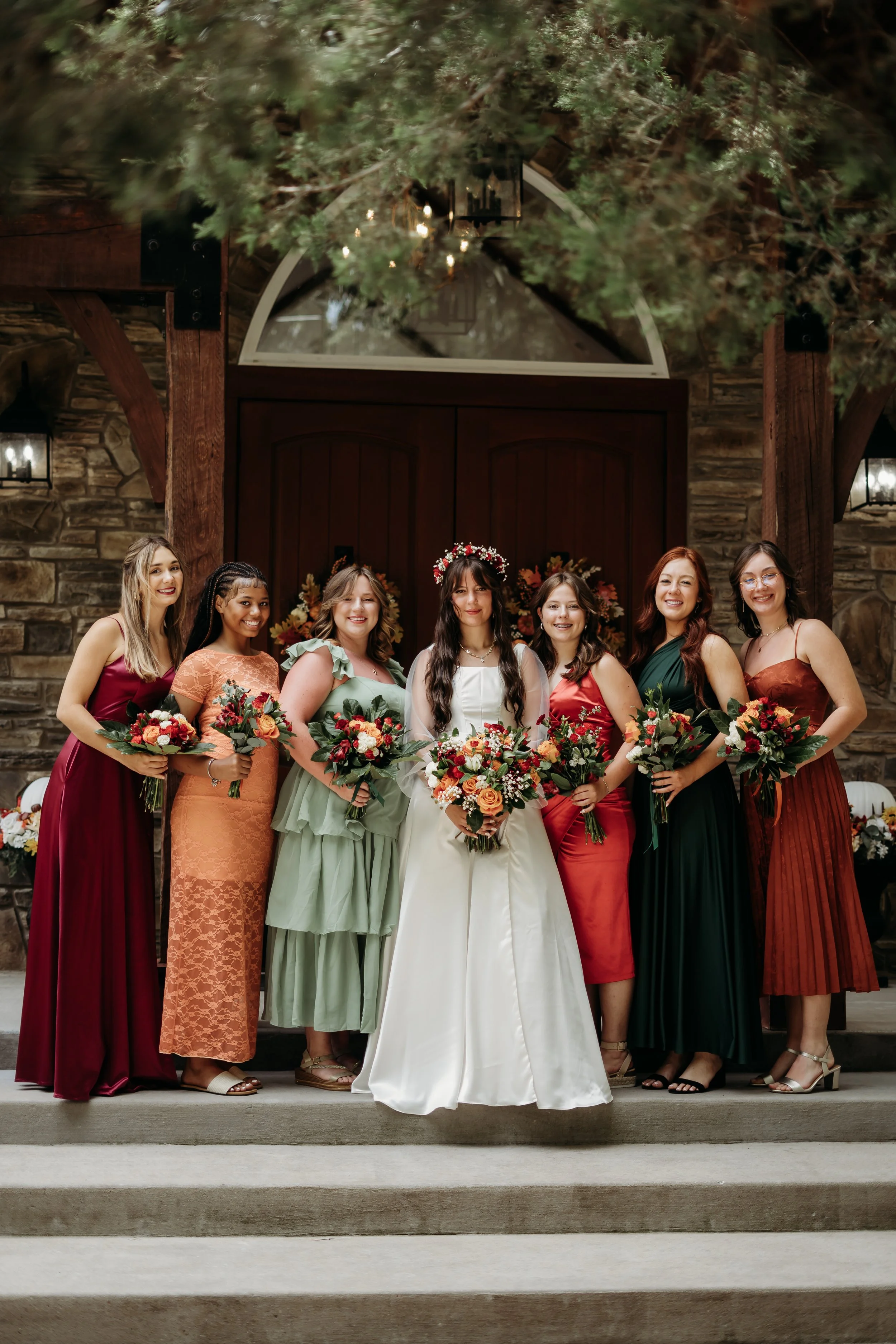 Group of eight women in colorful dresses standing on steps outside a rustic building, holding bouquets, celebrating a wedding.