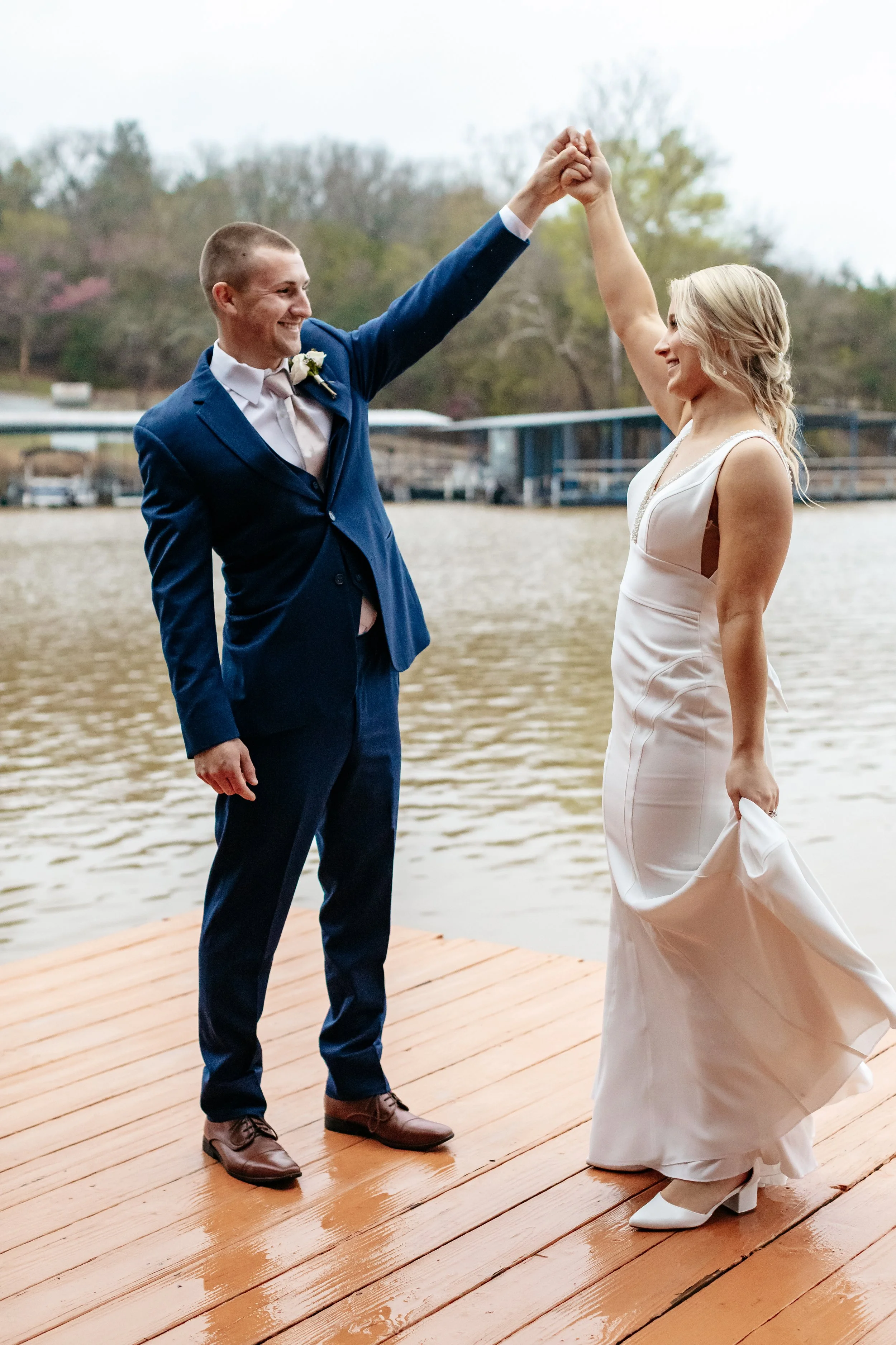 A couple in formal wedding attire dancing on a wooden dock by a lake with trees in the background.