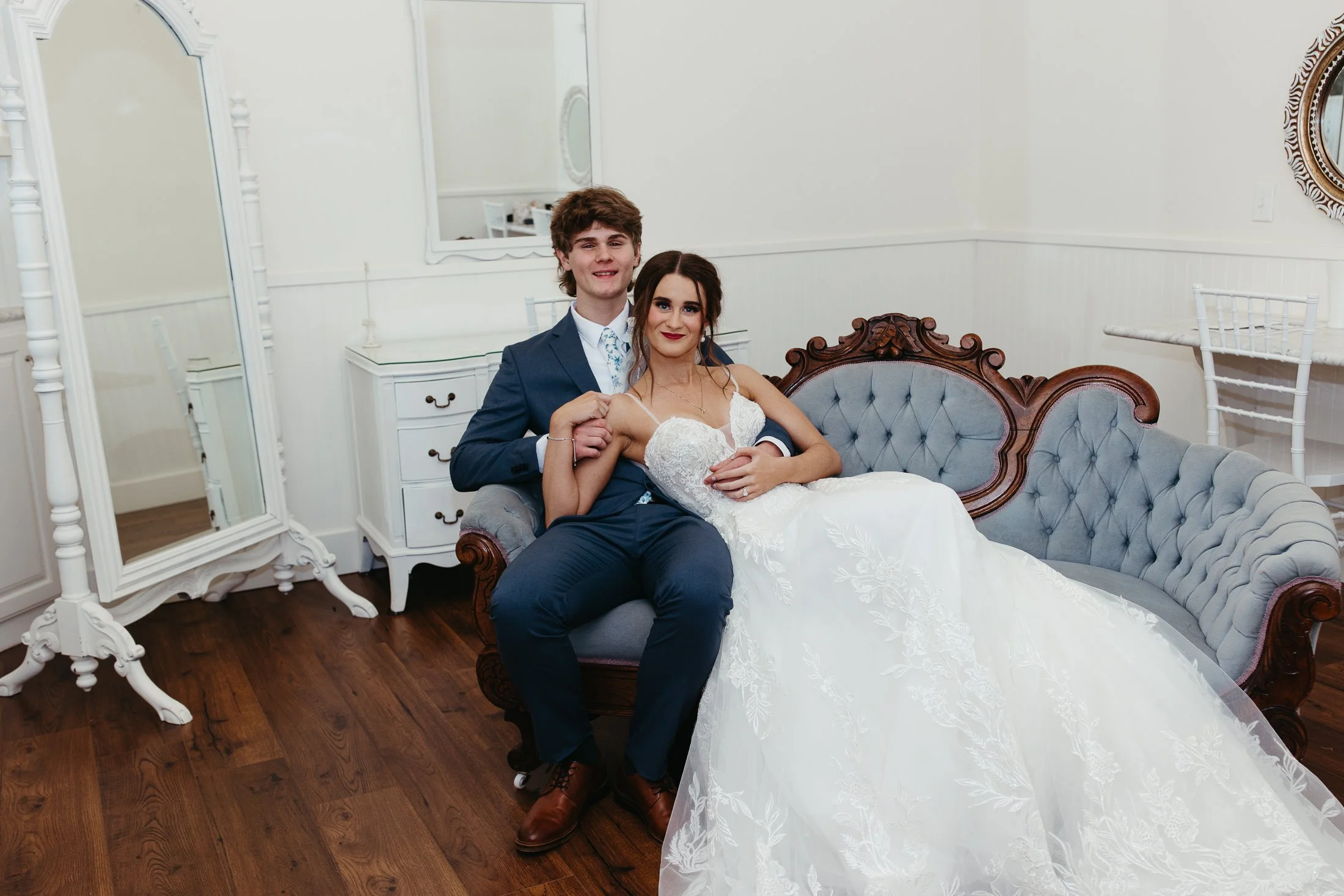 A young couple dressed in wedding attire sitting on a vintage gray sofa in a room with white walls, a large ornate mirror, a white dresser, and decorative chairs.