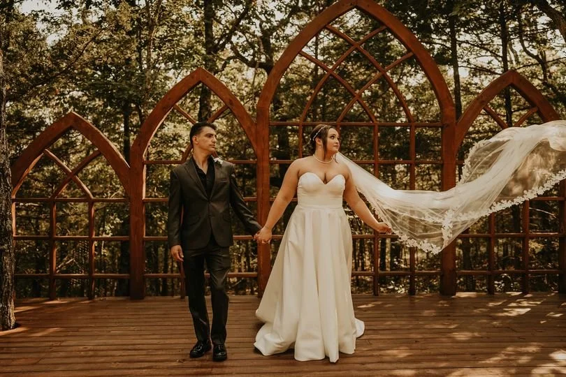 A bride and groom holding hands on a wooden platform in front of a backdrop of tall, wooden gothic-style arches surrounded by trees.