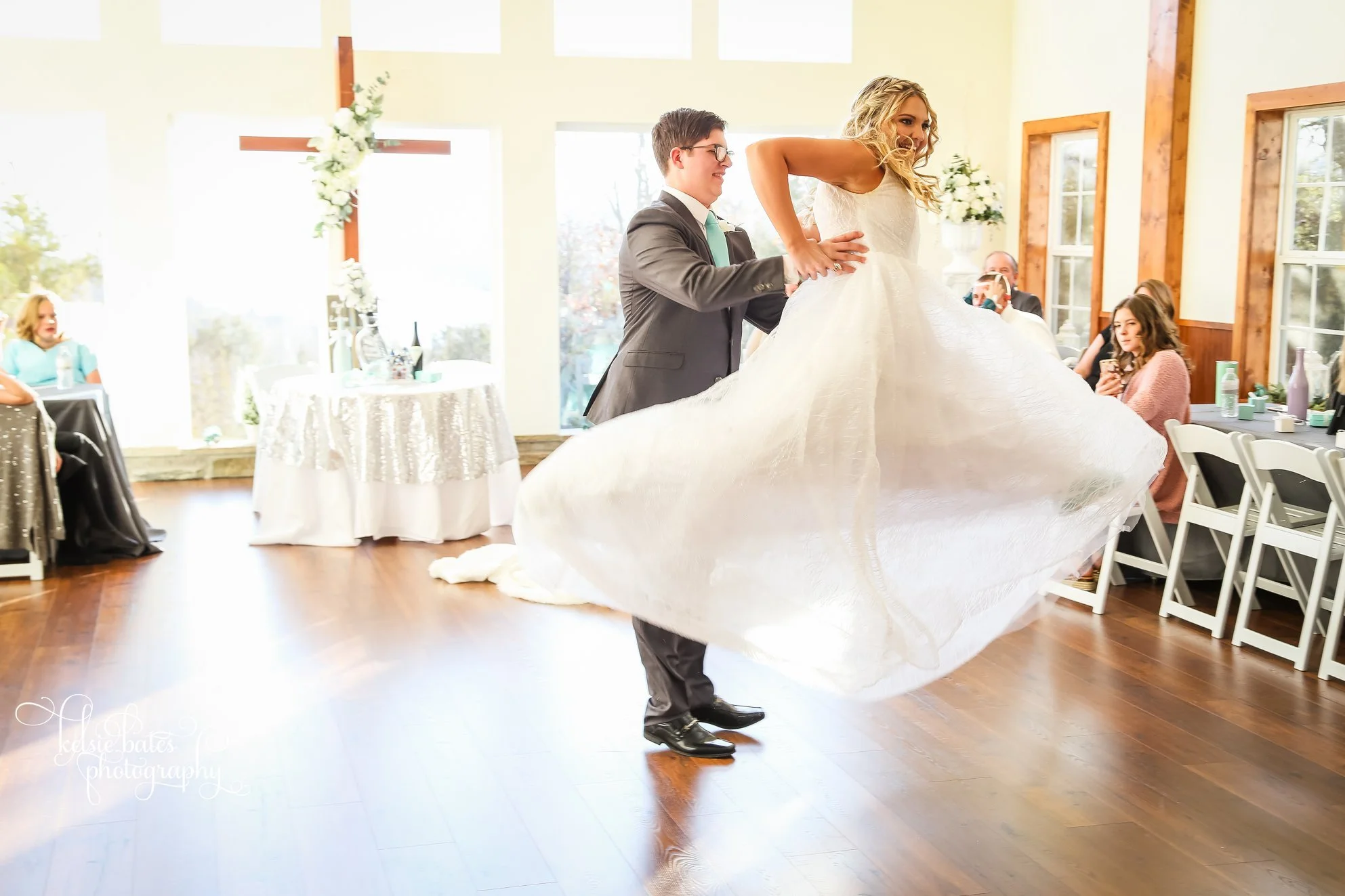 A bride and groom dancing at their wedding reception, with guests sitting at tables watching. The bride is wearing a white wedding gown and the groom is in a suit. The reception hall has large windows and wooden accents.