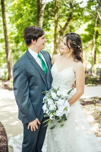 A newlywed couple standing close together outdoors, with the bride holding a bouquet of white flowers and both smiling at each other.
