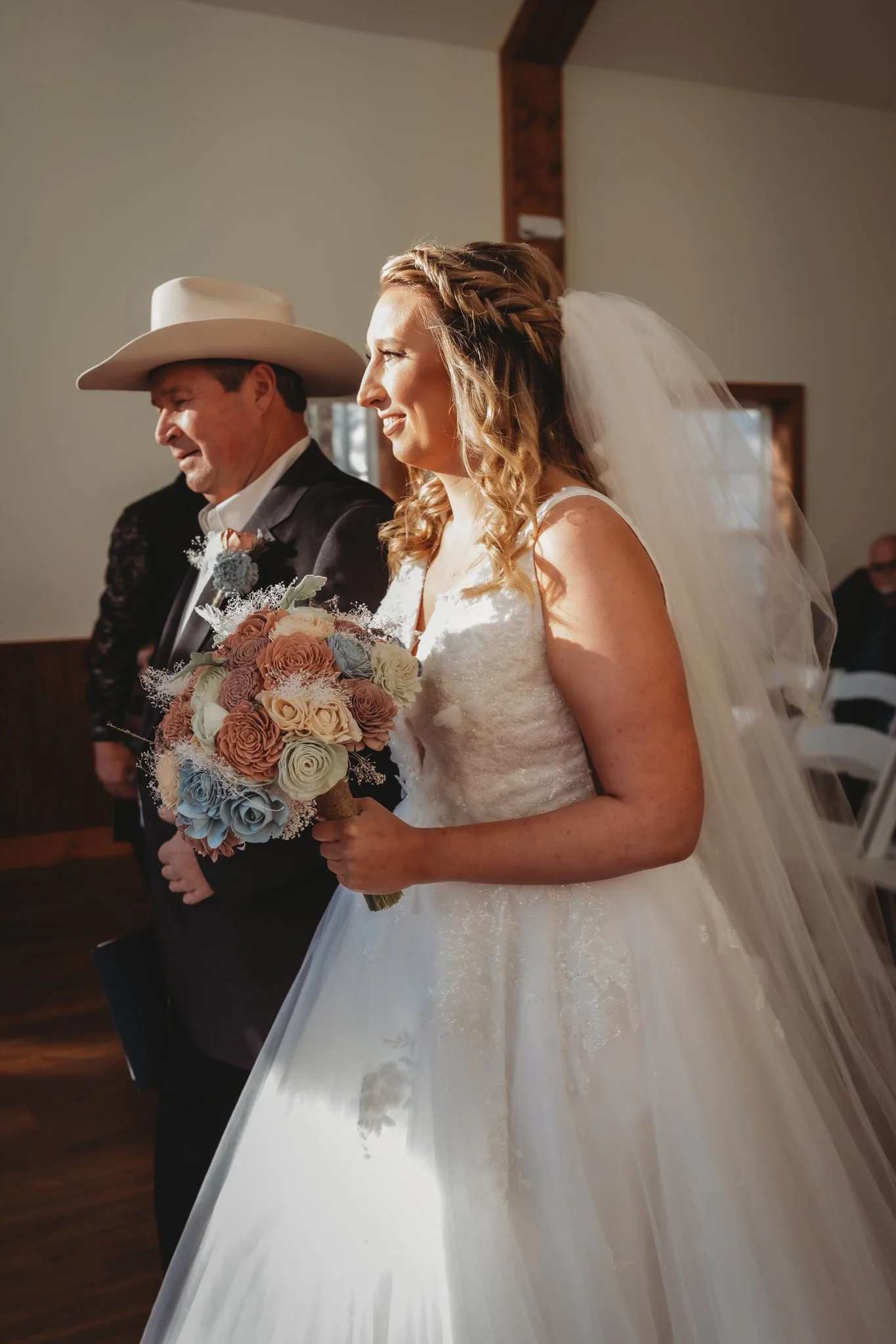 A bride with wavy blonde hair and a white wedding gown holding a bouquet of roses and other flowers, standing beside an older man wearing a large white cowboy hat and a dark suit, inside a church with wooden beams and cream-colored walls.