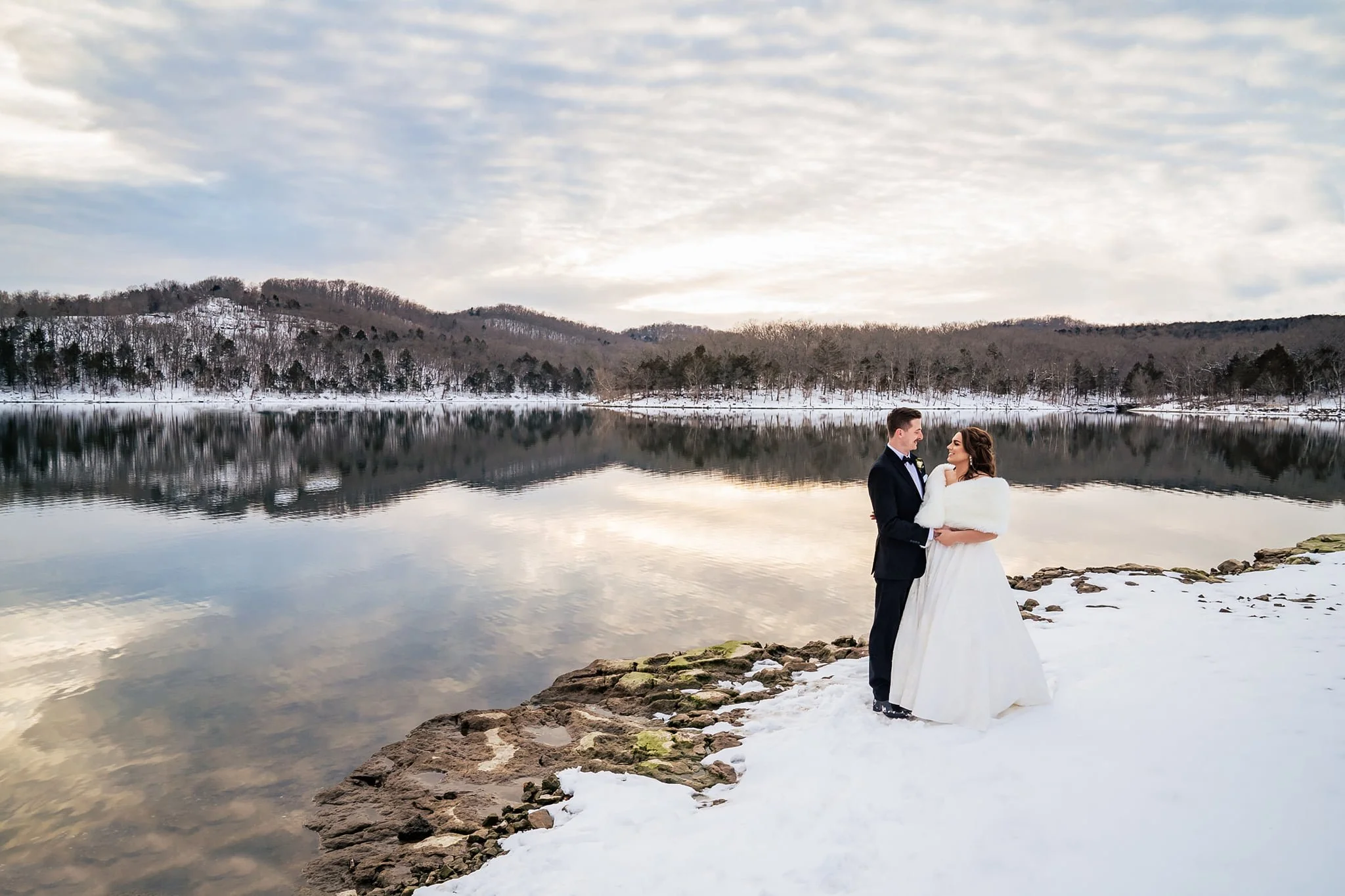 A bride and groom standing by a snowy lakeshore during winter, with snow-covered hills and trees in the background, and reflected cloudy sky in the calm lake, during daytime.
