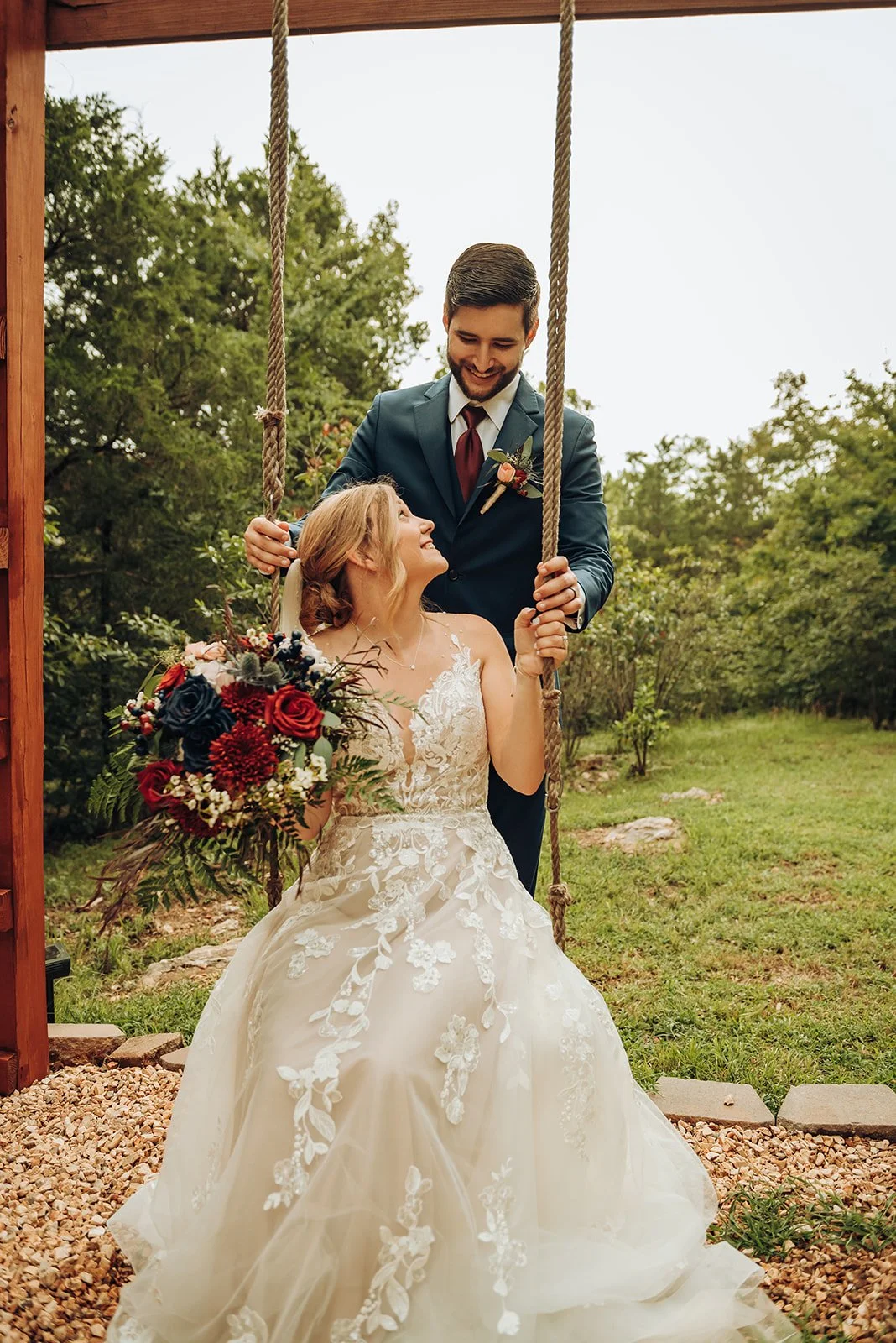 A bride and groom at their wedding outdoors, with the bride sitting on a swing holding a bouquet of flowers, and the groom standing next to her, smiling.