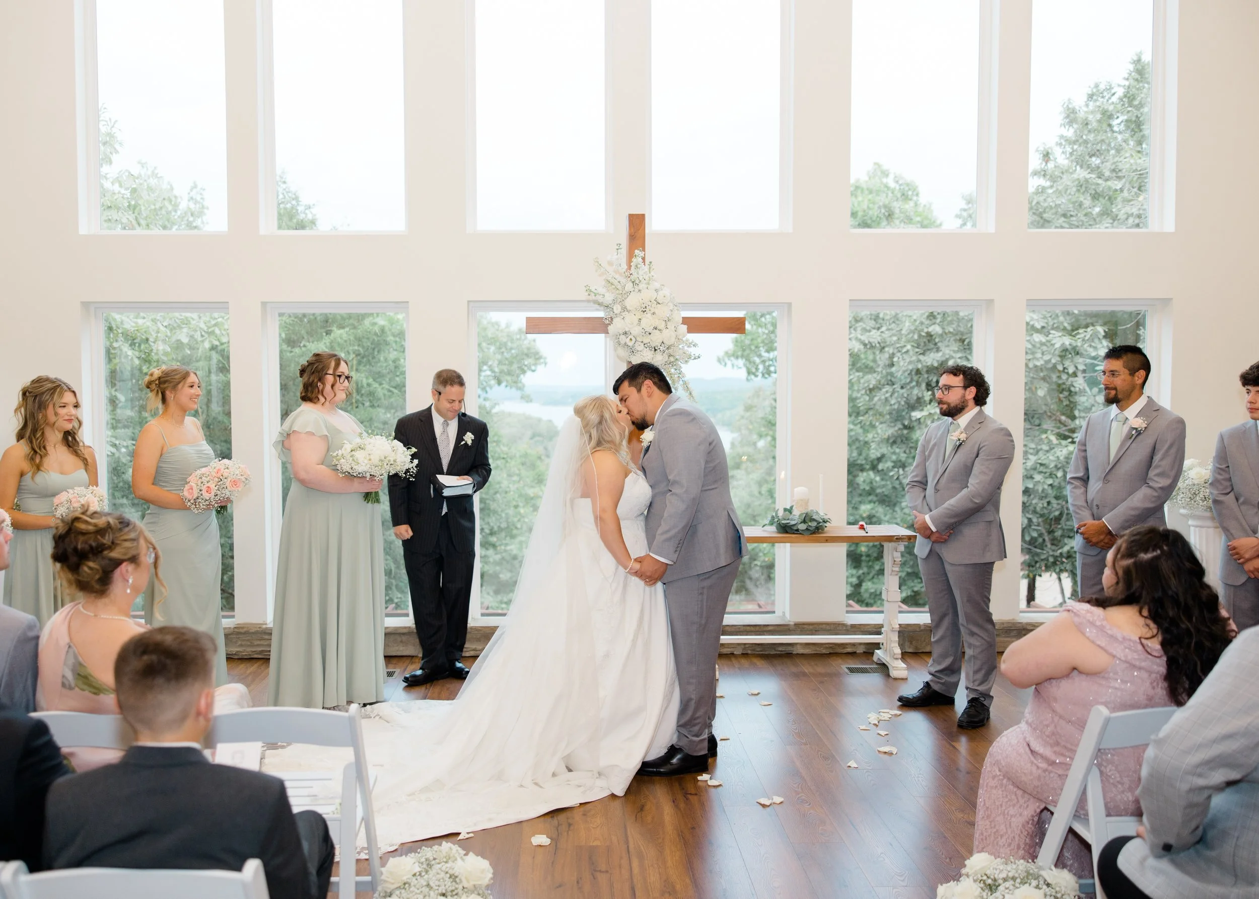 A wedding ceremony inside a church with large windows overlooking trees. The bride and groom are kissing at the altar, surrounded by bridesmaids and groomsmen. The church has a wooden cross decorated with flowers behind the couple.