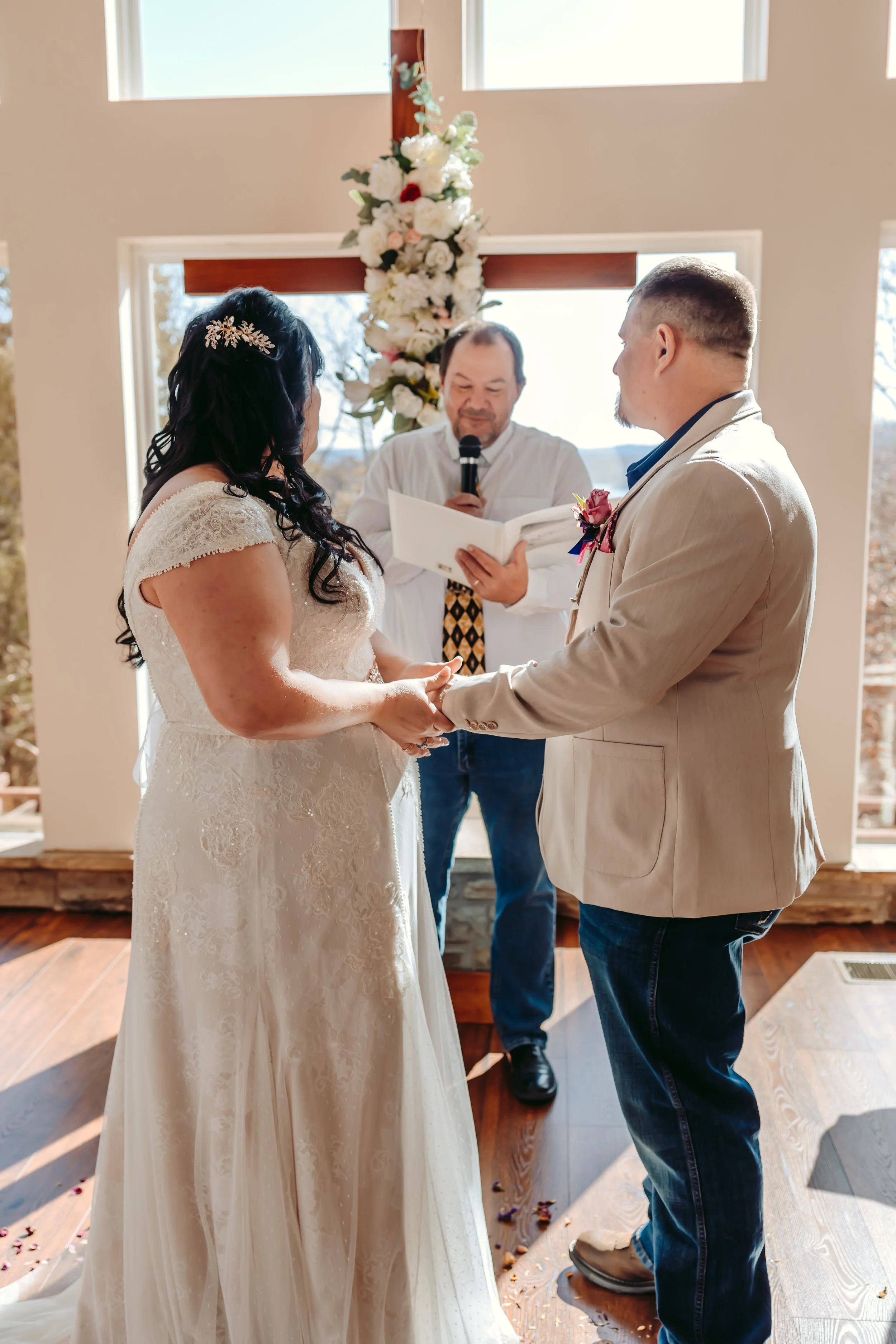 A bride and groom exchange vows during a wedding ceremony inside a decorated venue with a wooden cross and floral arrangement behind them. An officiant is reading from a paper, and the couple holds hands.