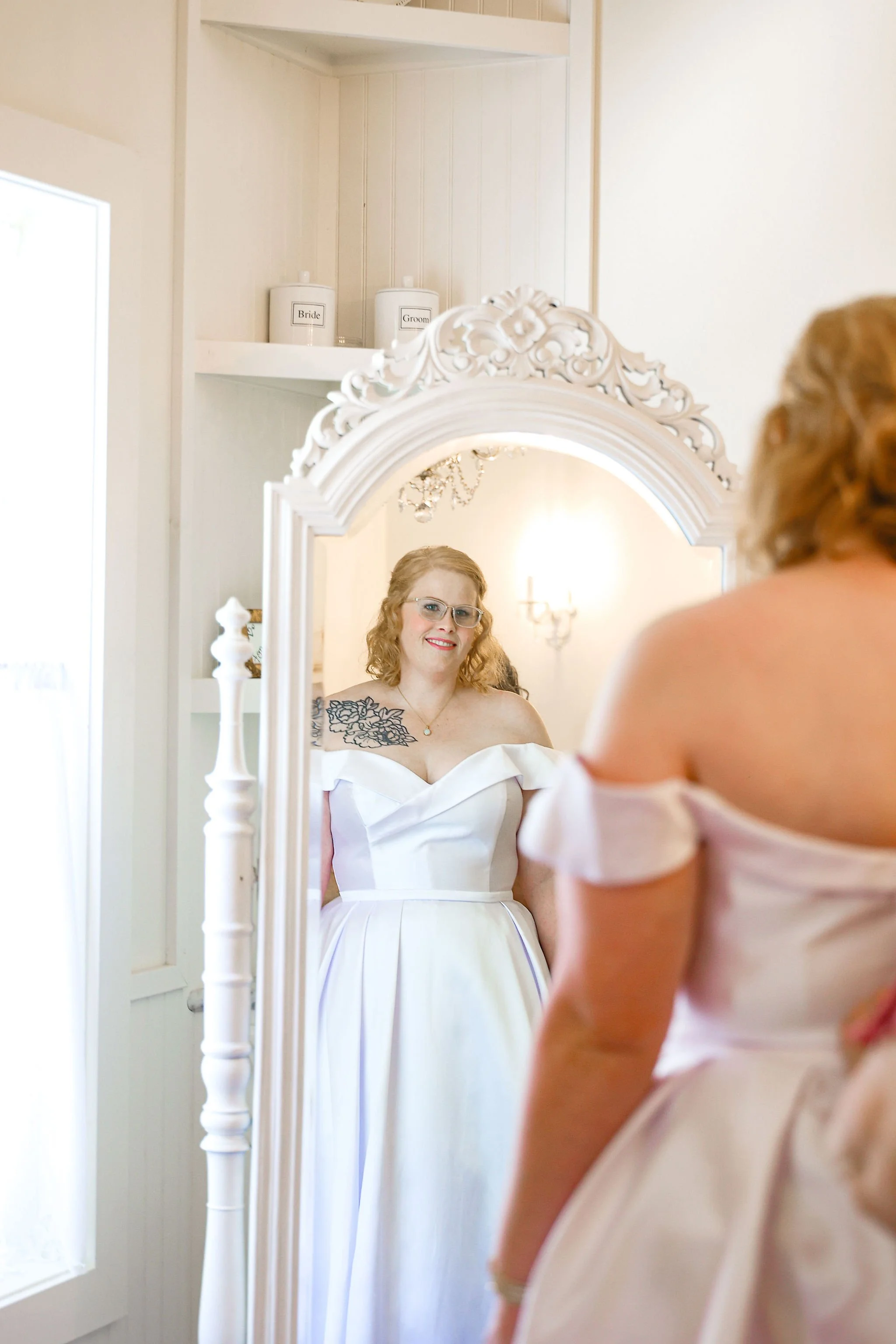 A woman in a wedding dress looking at herself in a mirror, with a young girl in a pink dress partially visible in the foreground, in a bright, elegant room.