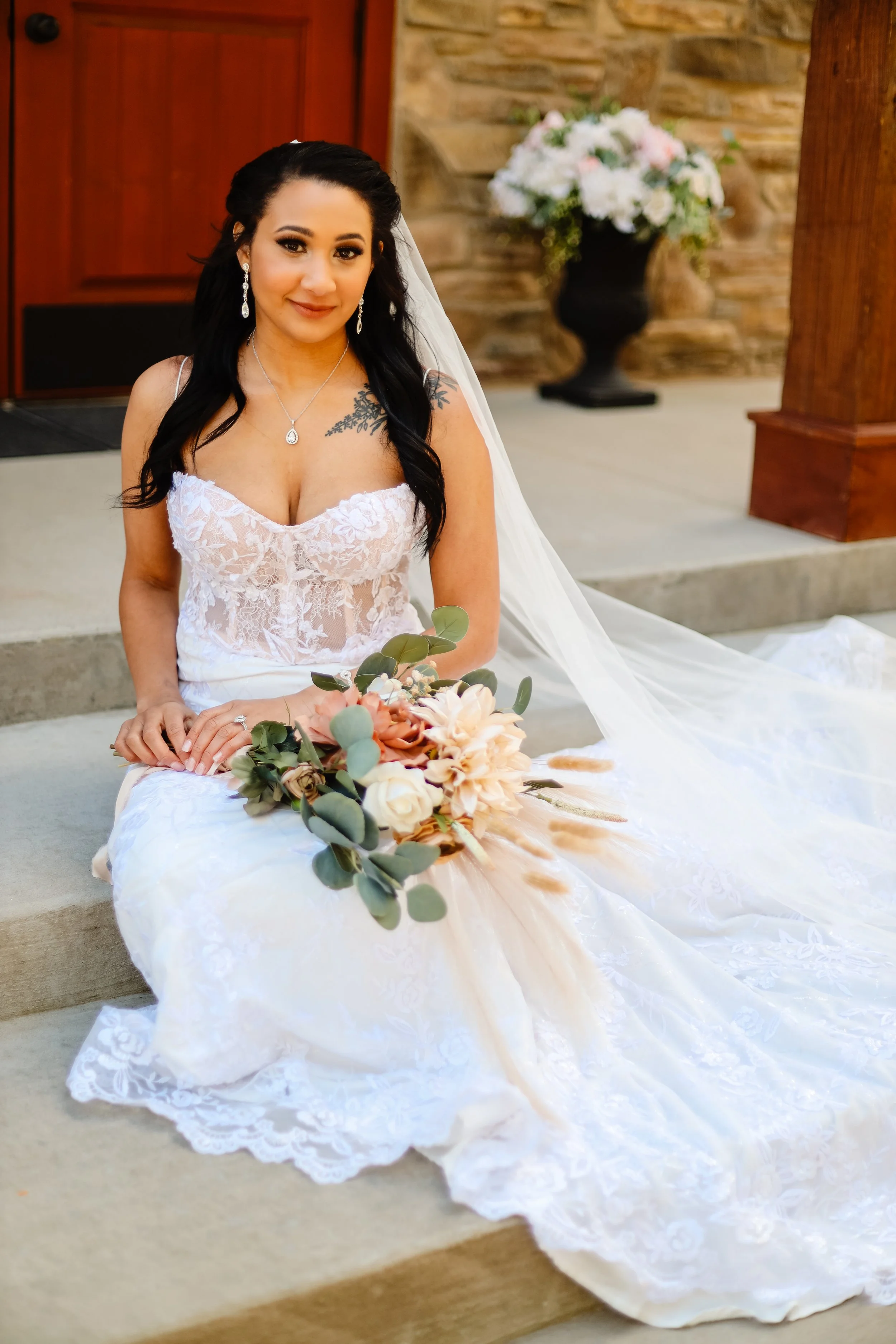A bride in a white lace wedding dress with a necklace, earrings, and veil, sitting on a stone step holding a bouquet of pink, white, and beige flowers with greenery, in front of a rustic stone wall with a black vase of white and pink flowers in the b