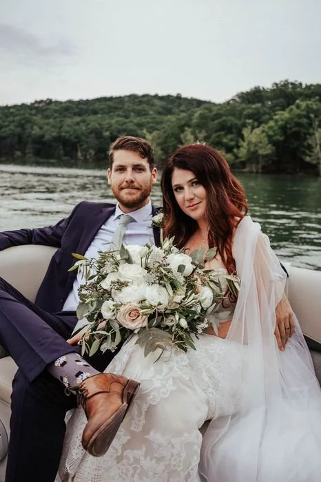 A newlywed couple sitting together on a boat, with water and green trees in the background, the bride holding a large bouquet of white flowers.
