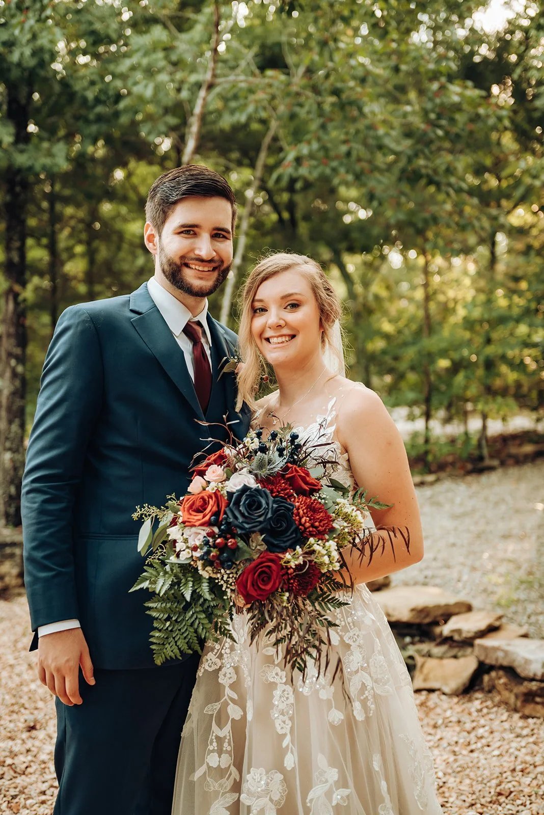 A newlywed couple smiling outdoors, the groom in a dark suit and the bride in a lace wedding dress holding a bouquet of red, navy, and white flowers.