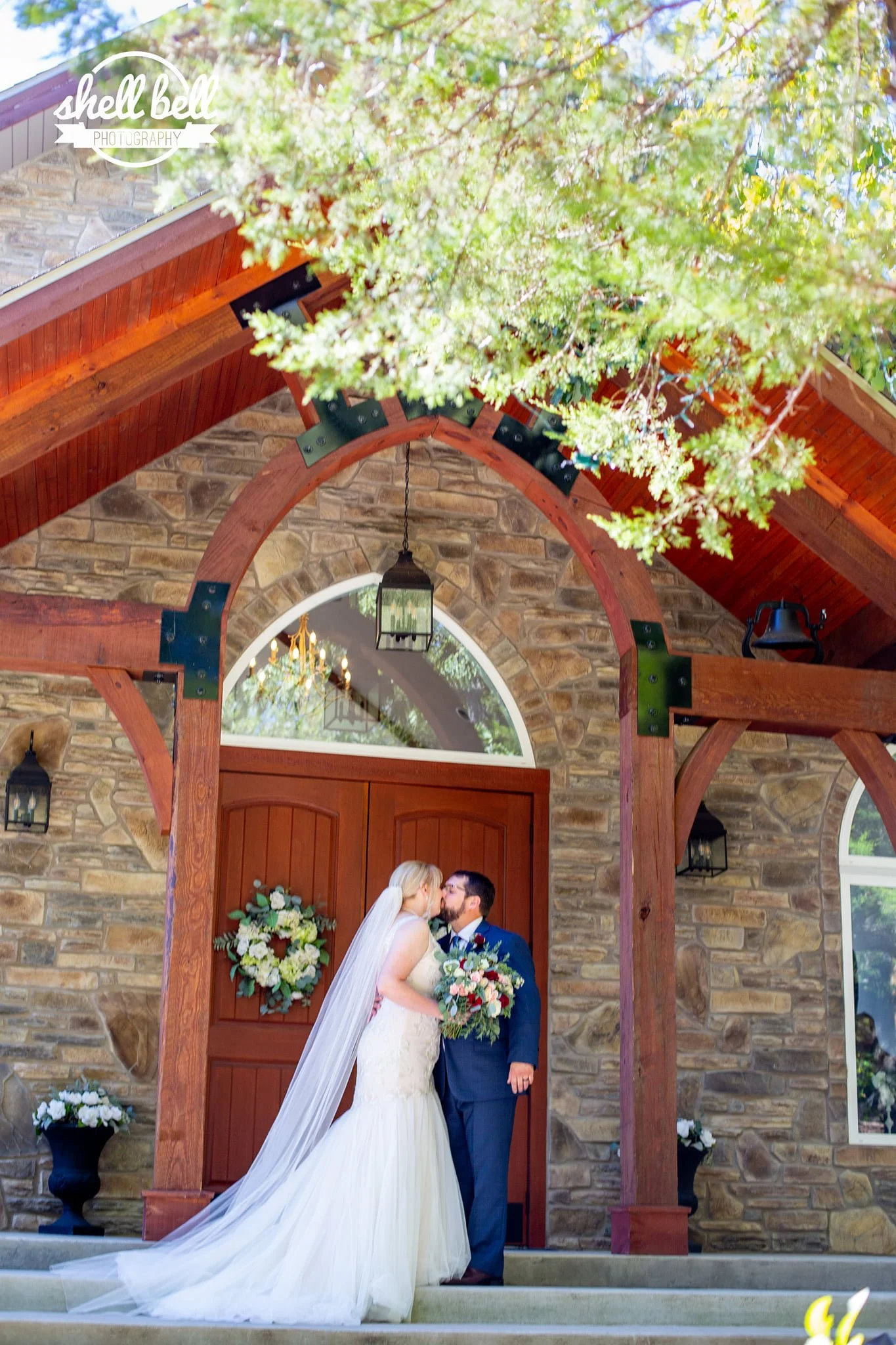 A bride and groom kiss under an arched wooden structure decorated with floral arrangements during a wedding ceremony outside a stone building.