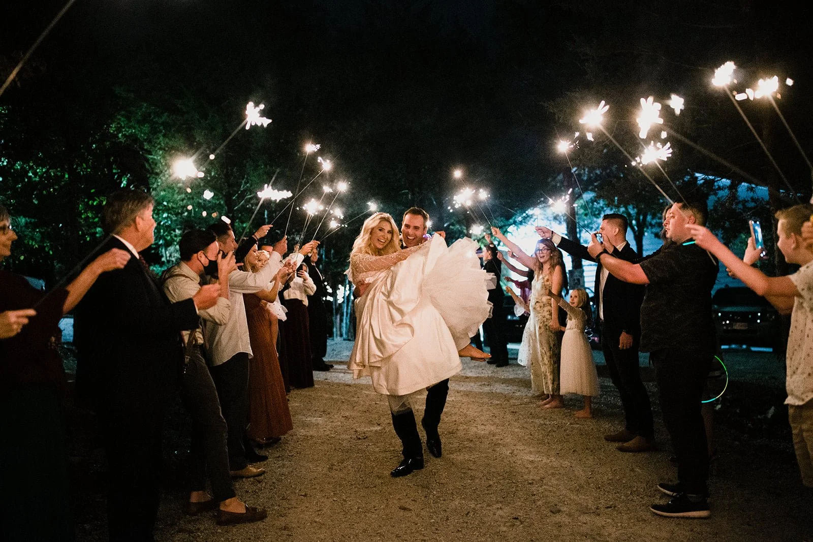 A bride and groom walking and smiling through a line of guests holding sparklers at night during a celebration.