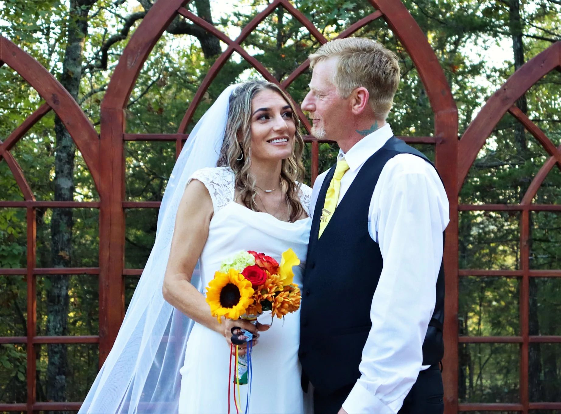 A bride and groom standing close together outdoors, with a red garden arch and trees in the background. The bride is smiling, holding a bouquet of sunflowers, roses, and other flowers. The groom is looking at her, smiling, wearing a vest and a yellow tie.