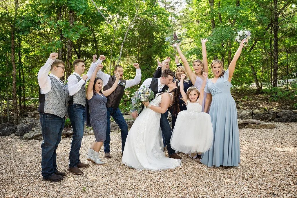 Group of people celebrating wedding outdoors with trees in the background, including a bride and groom, bridesmaids, groomsmen, and a young girl in a white dress.